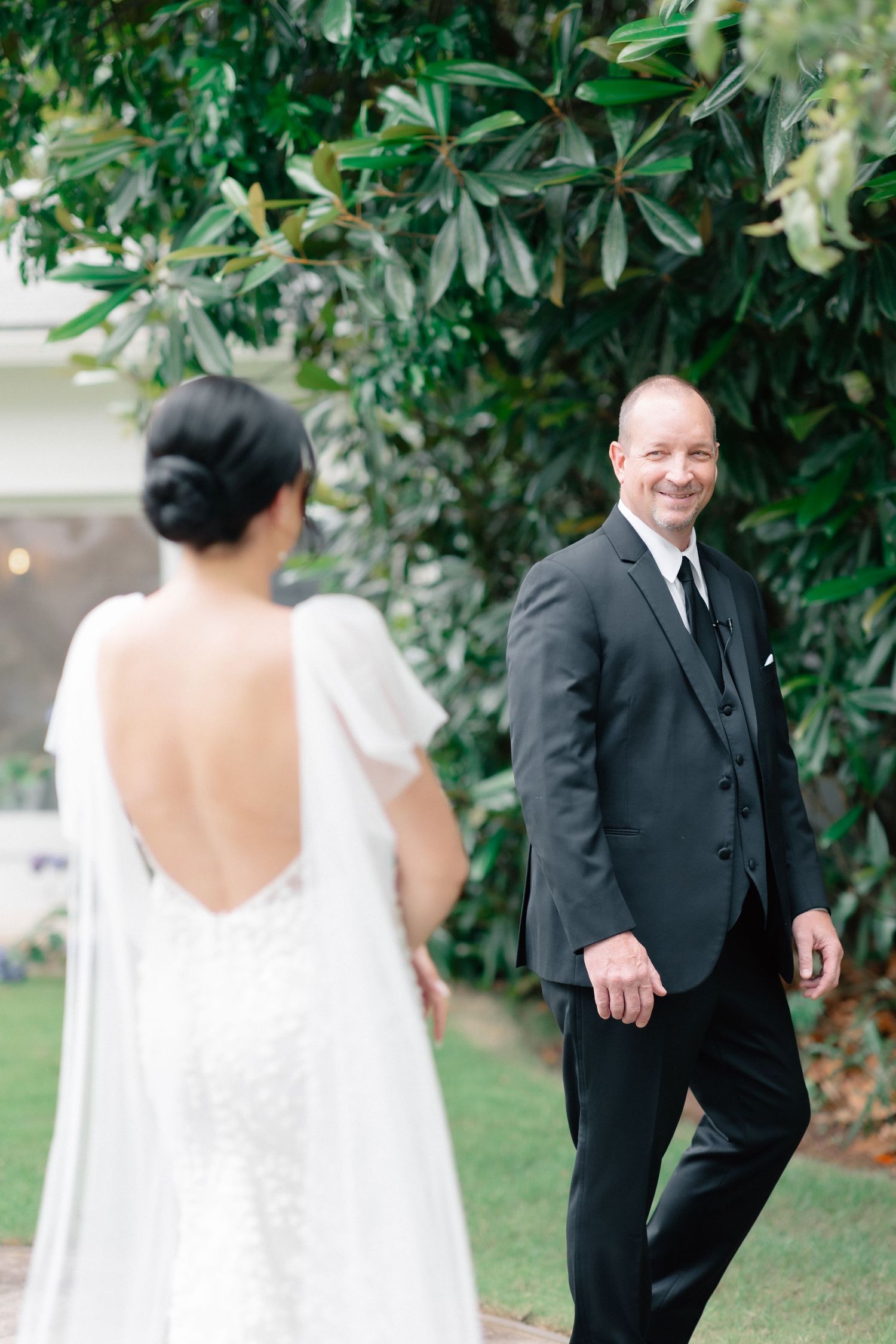 Groom in black suit smiling at bride in backless lace wedding dress during first look outdoors.
