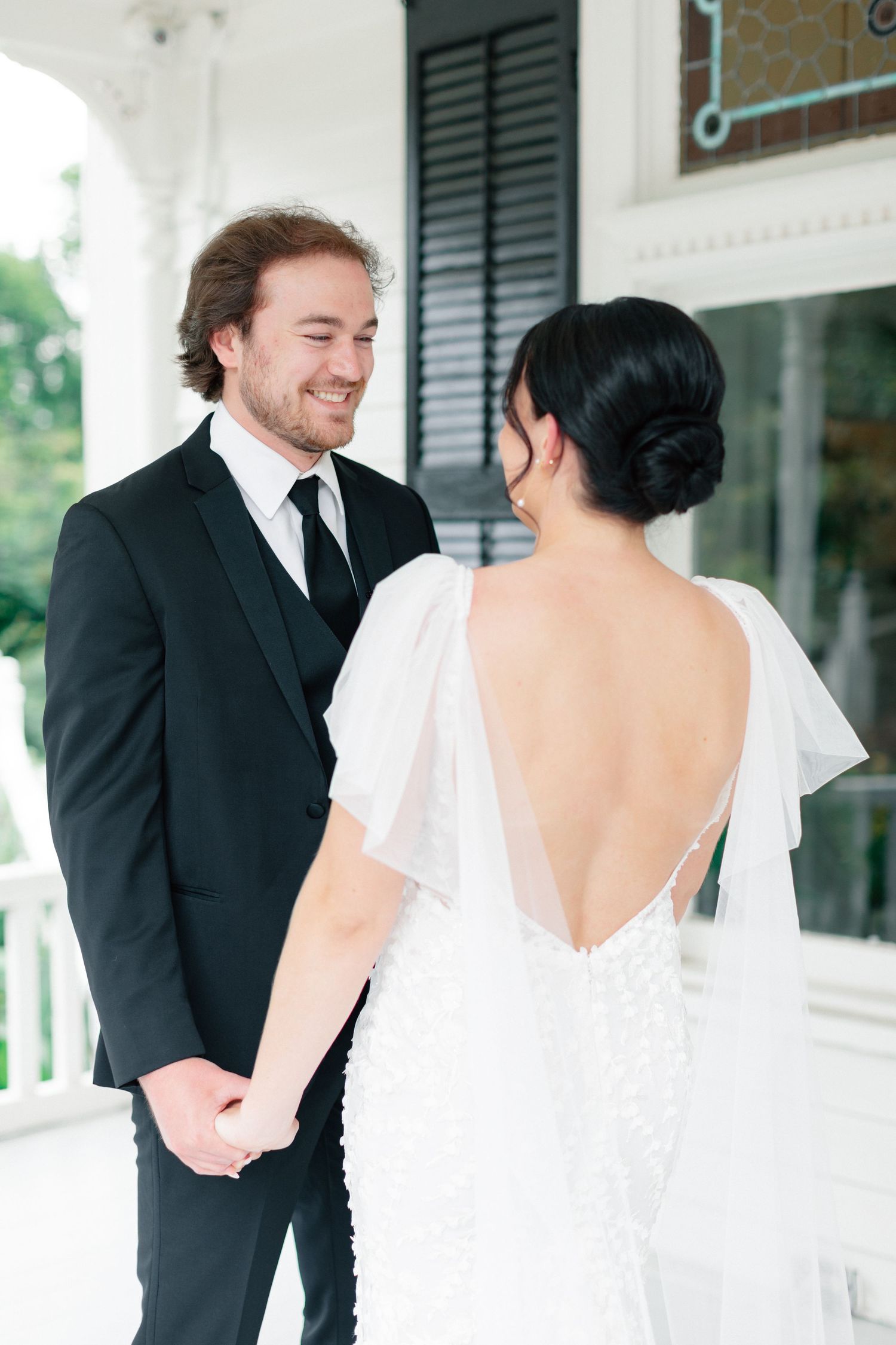 Groom in black suit smiles at bride in backless beaded wedding gown during first look on white porch.