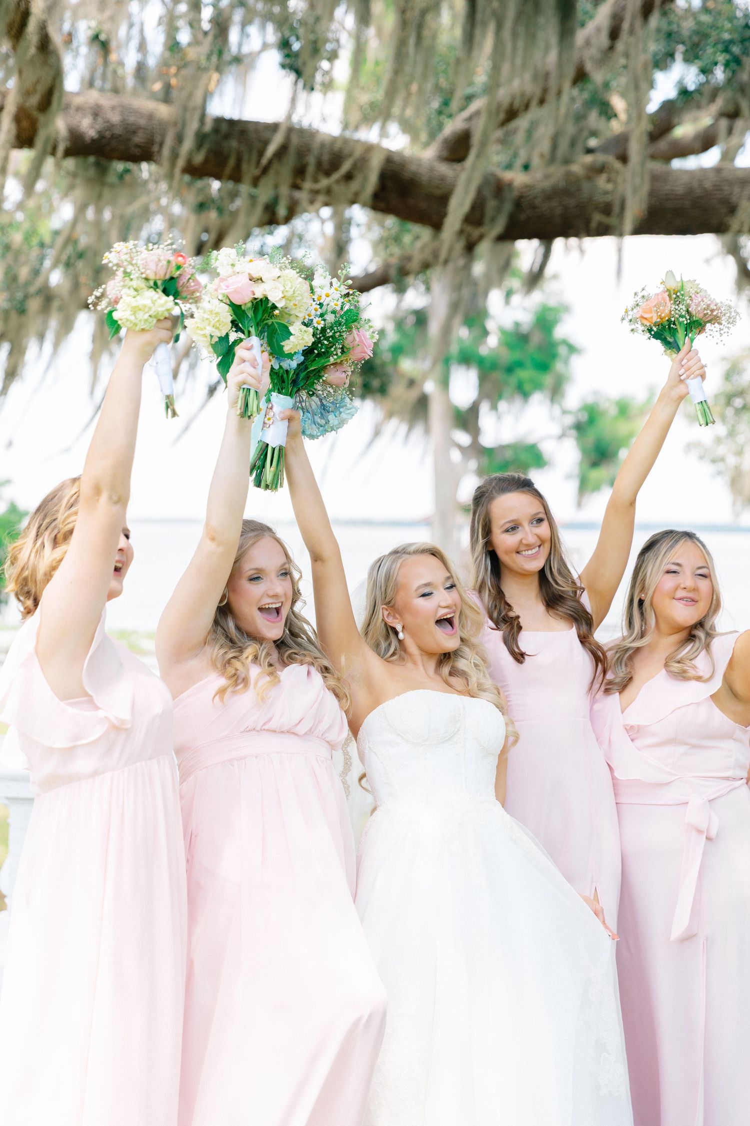 Bride in white strapless gown celebrates with four bridesmaids in blush pink dresses, raising floral bouquets outdoors, wedding at Bella Cos
