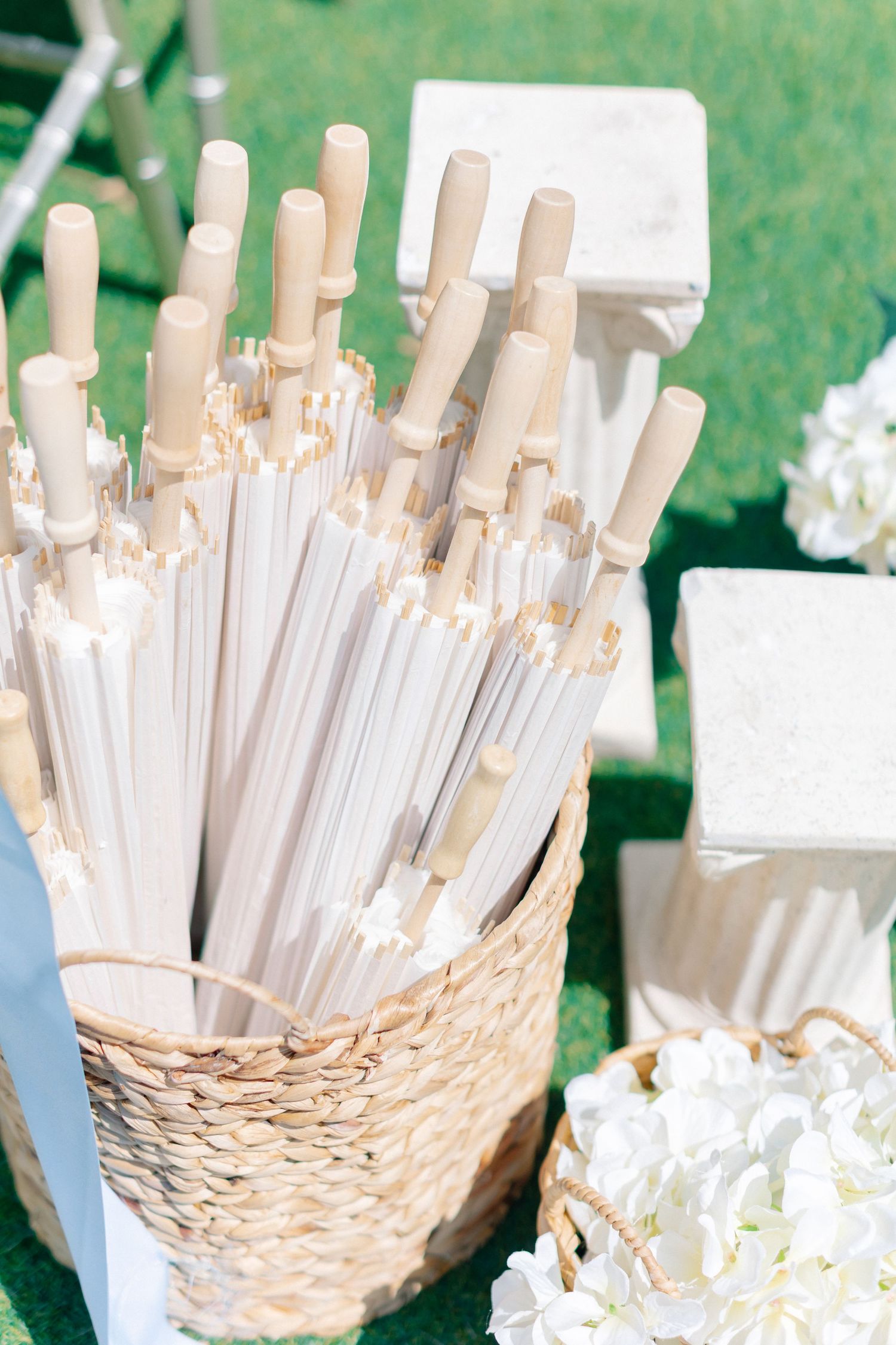 White parasols in a woven basket displayed outdoors at a wedding with white hydrangeas and gift boxes nearby, wedding at Bella Cosa