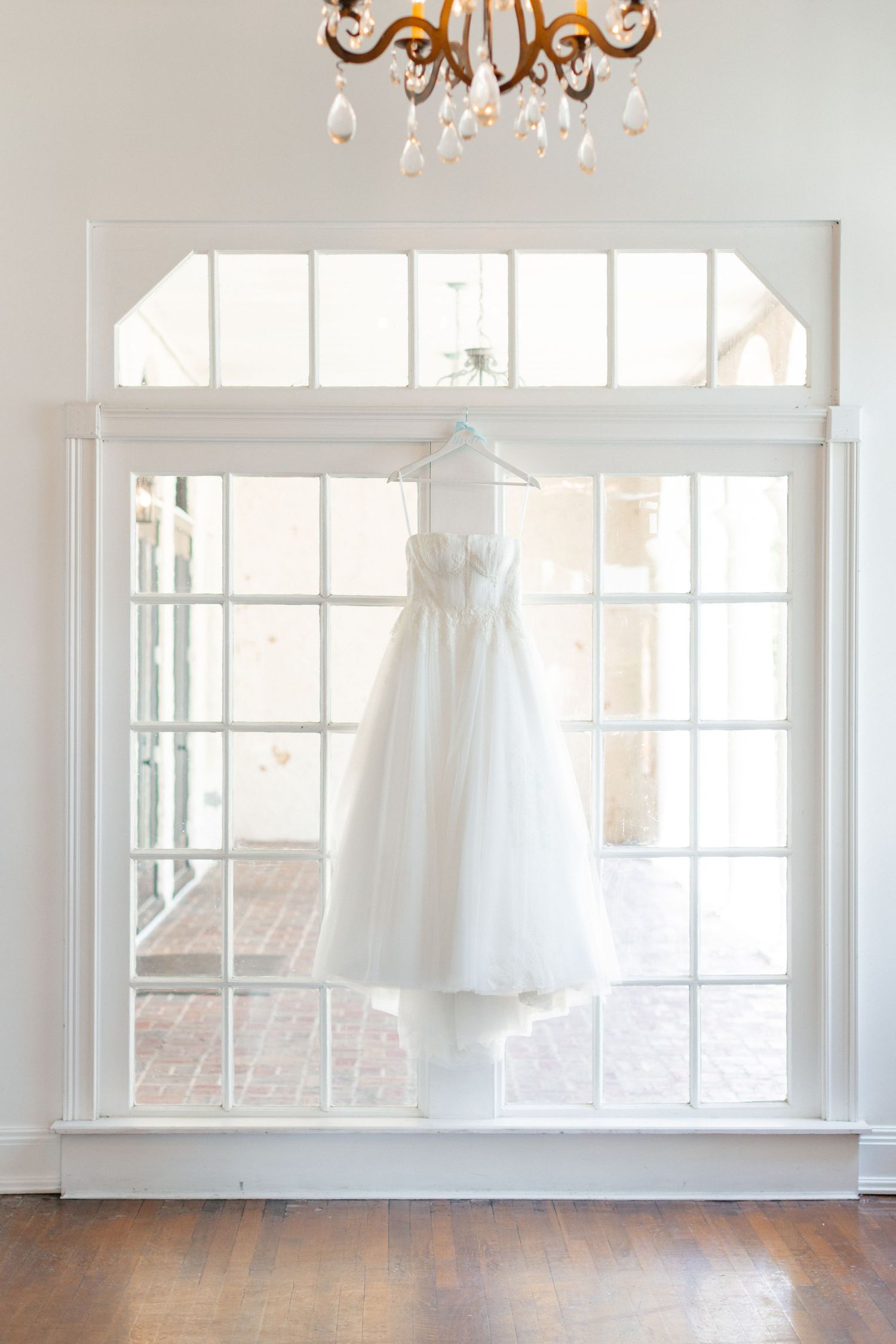 A white bridal gown hanging on a hanger in front of a large arched window with a crystal chandelier above, wedding at Bella Cosa