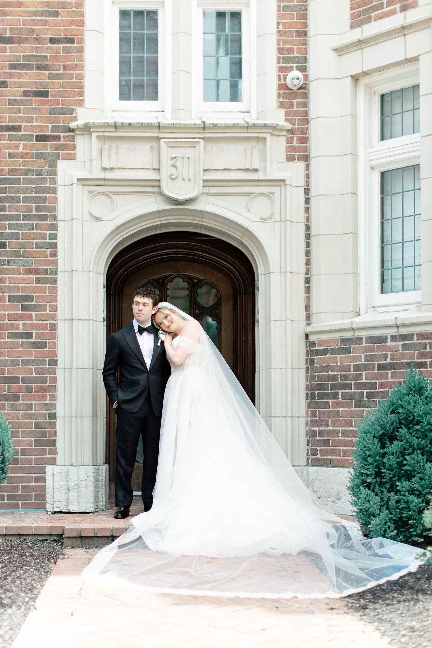 Bride and groom posing in front of elegant brick building arched doorway, bride's long veil flowing gracefully on ground.