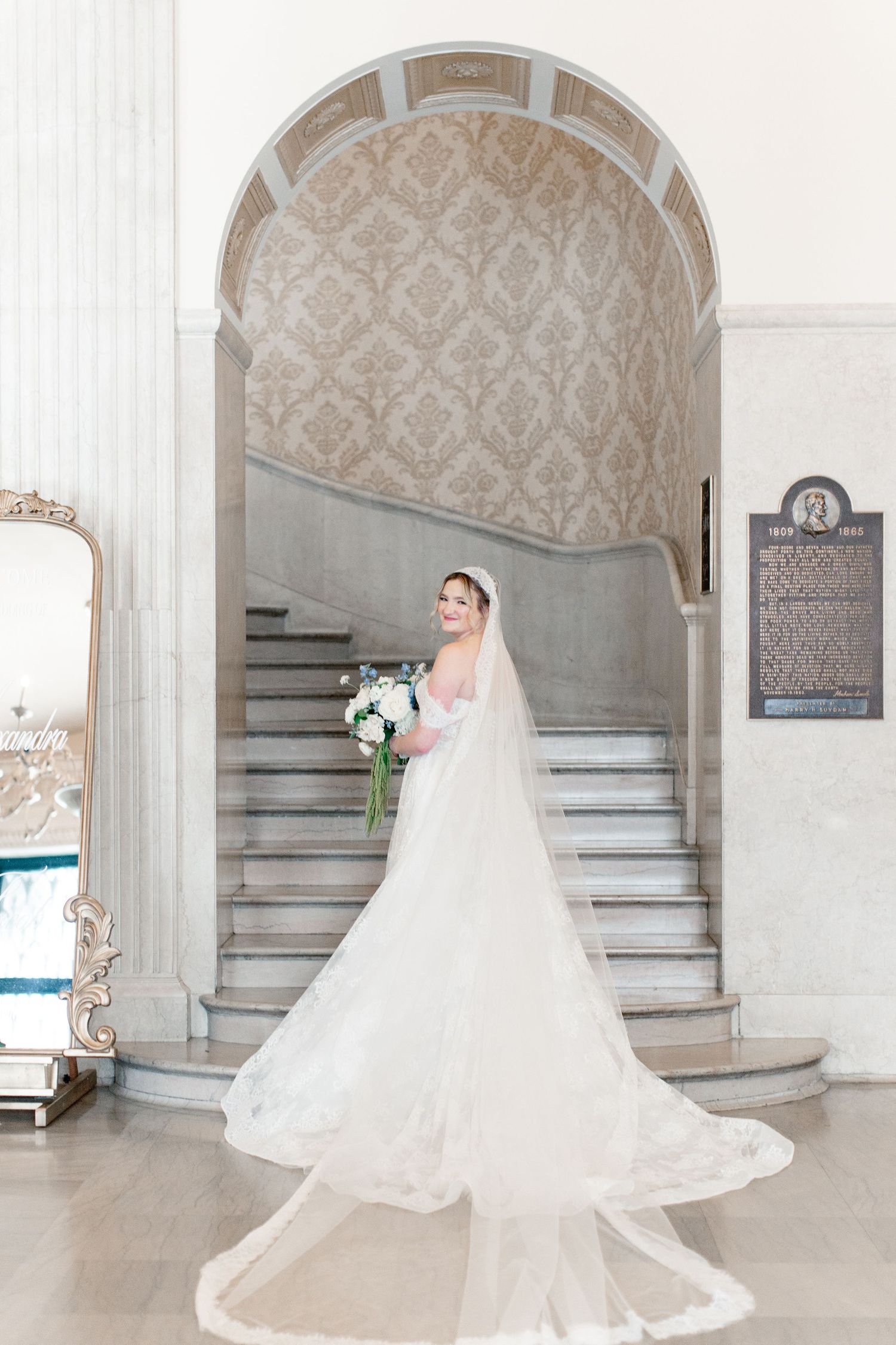 Bride in a flowing white gown with long veil holds bouquet on grand marble staircase in elegant venue.