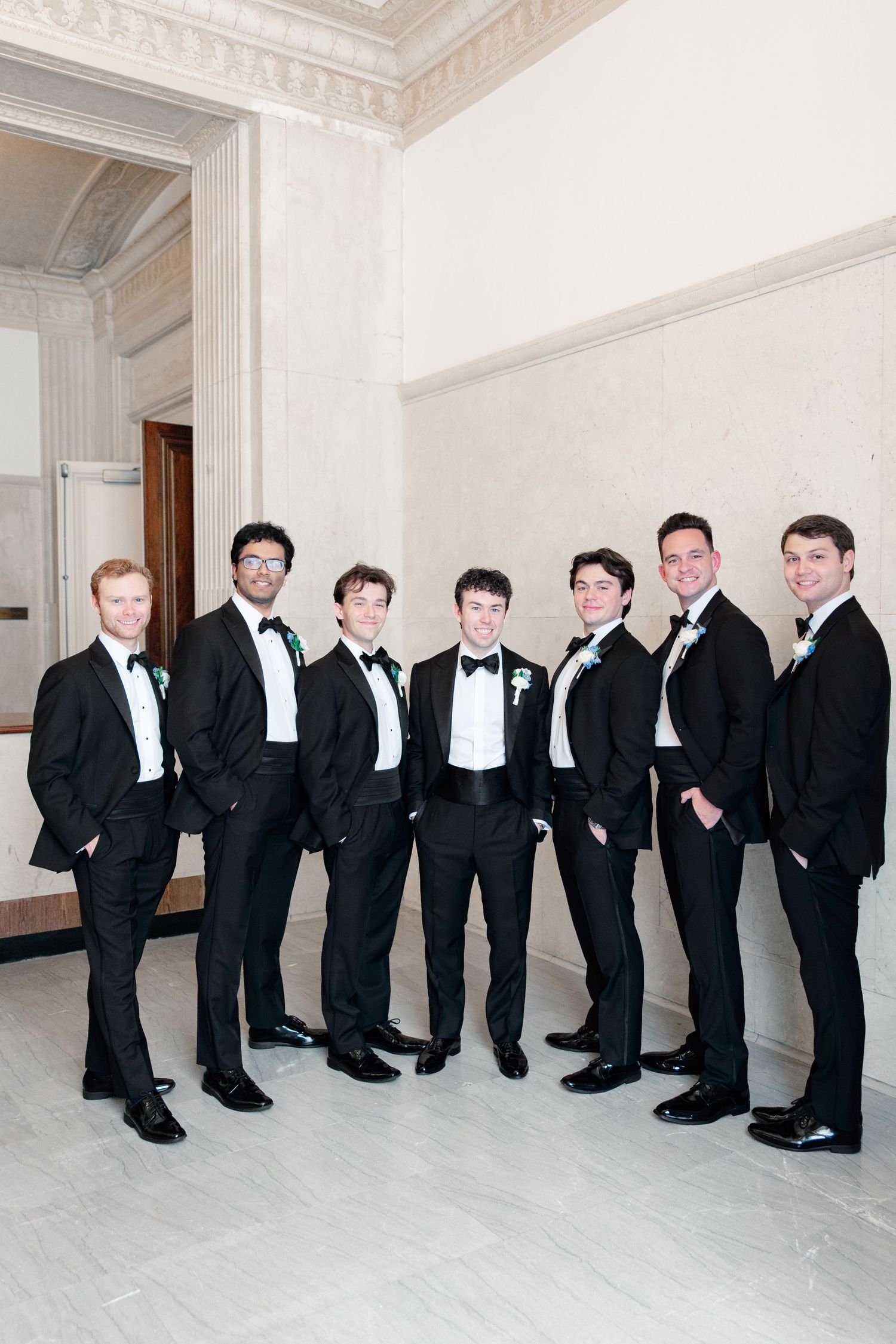Seven groomsmen in black tuxedos with bow ties and boutonnieres posing together in an elegant white marble venue.