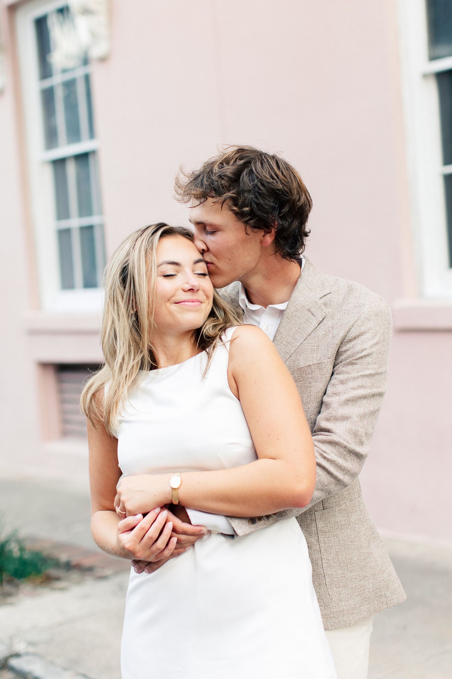 Man in tan blazer tenderly kissing woman on the temple as she smiles with closed eyes in front of pink building.