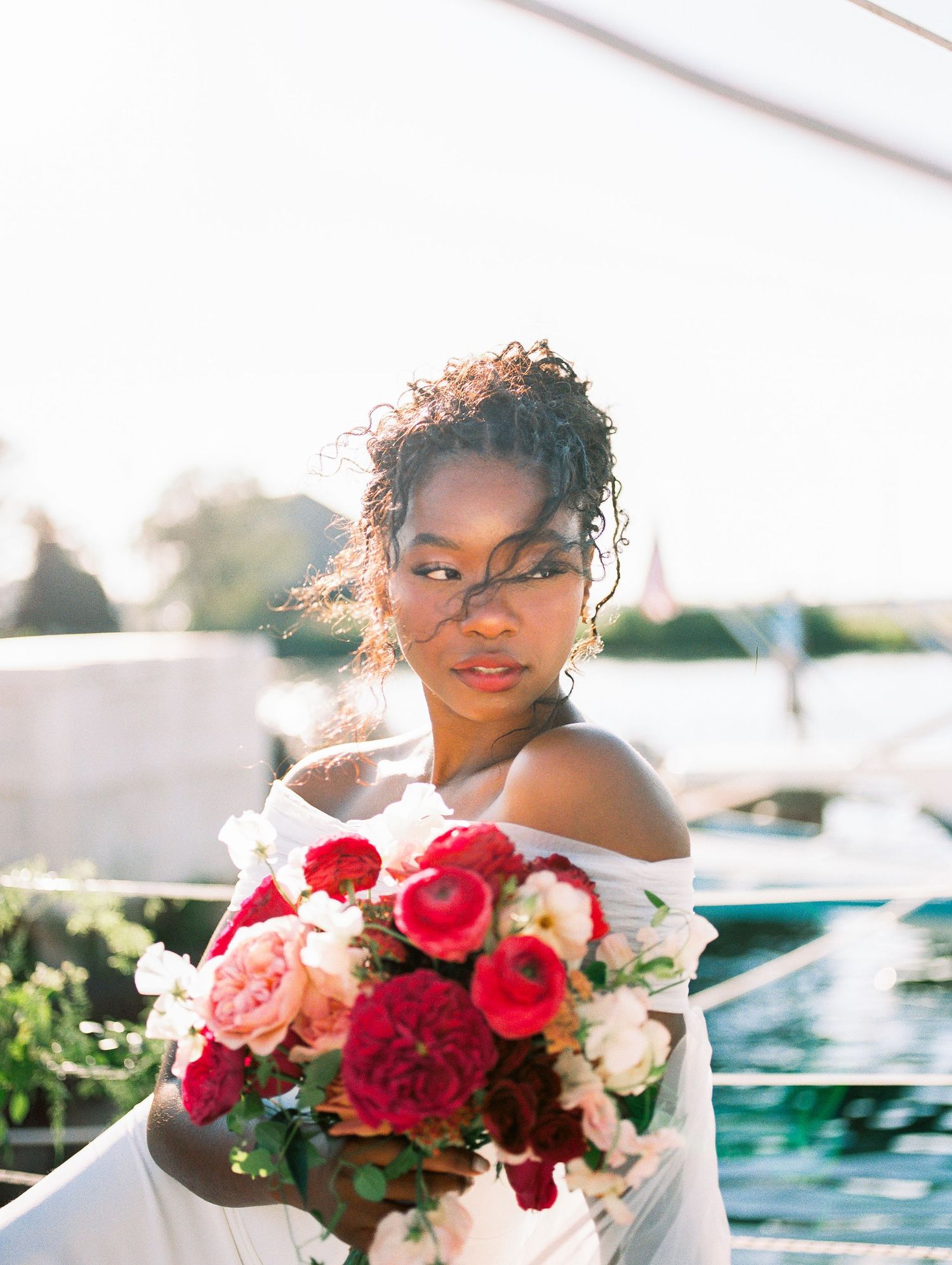 Beautiful Black bride holds a vibrant red and pink bouquet on a sunlit sailboat.
