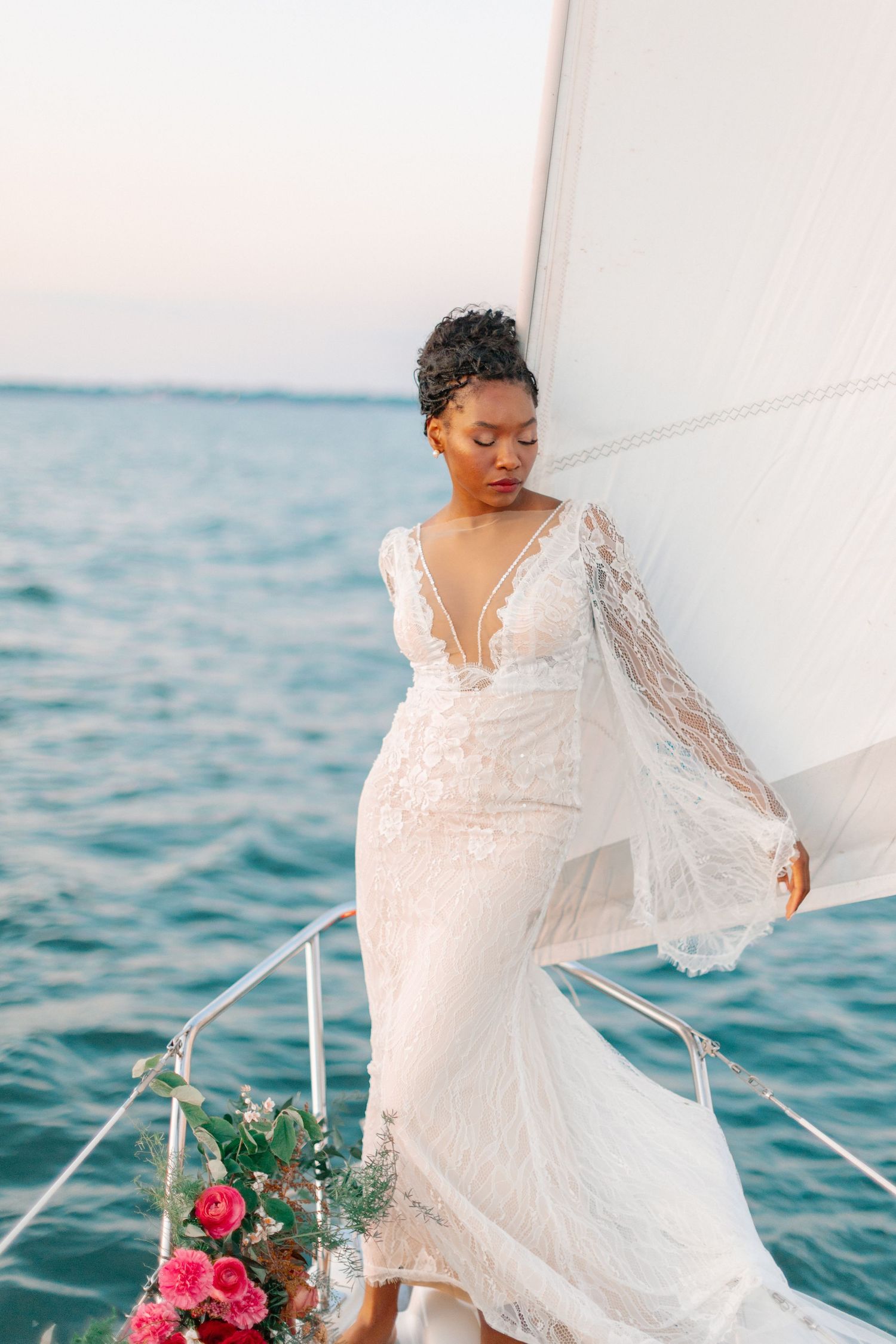 Elegant bride in lace gown on sailboat with ocean backdrop and pink floral bouquet.