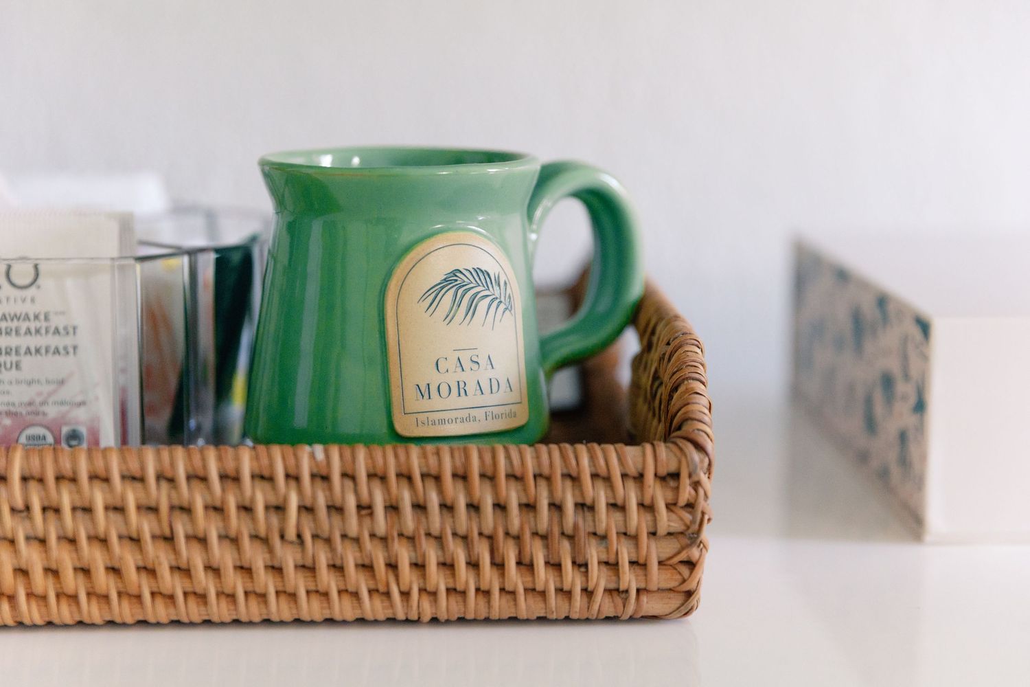 Green Casa Morada branded ceramic mug in a woven rattan tray with tea packets on a white surface.