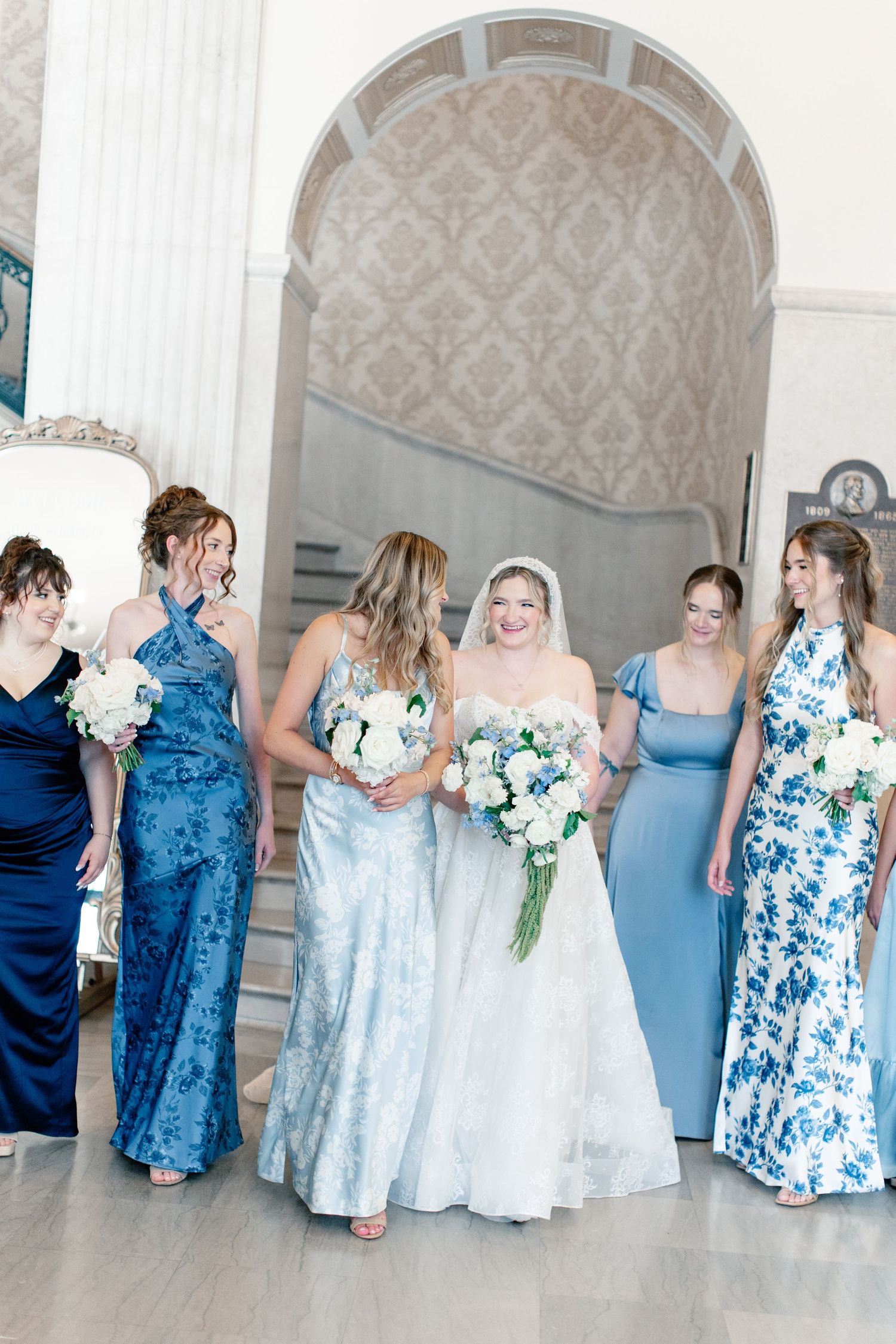 Bride in white gown with bridesmaids in blue dresses holding bouquets, laughing together in elegant venue.
