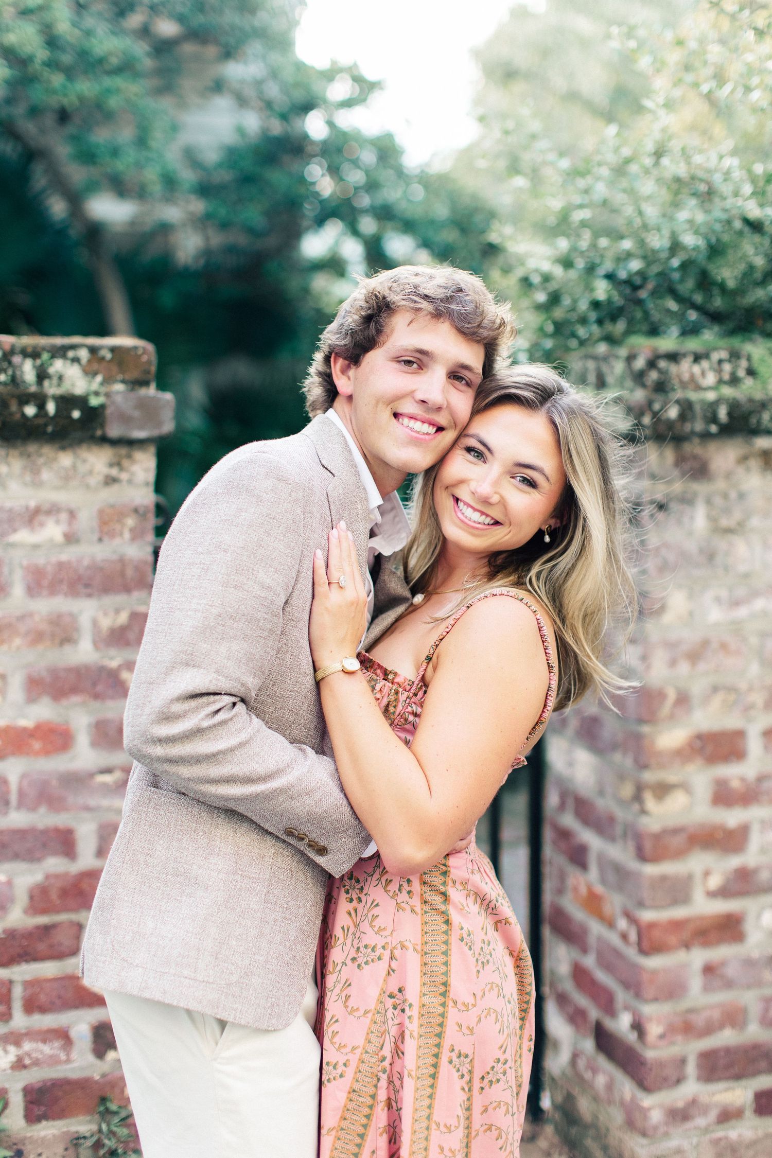 Smiling couple embracing near a brick wall, woman in pink floral dress showing engagement ring, man in tan blazer.