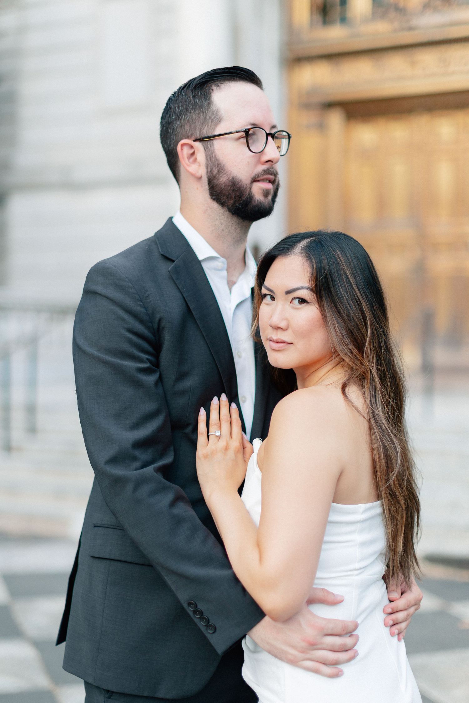 Engaged couple embracing outside ornate stone building, woman in white dress.