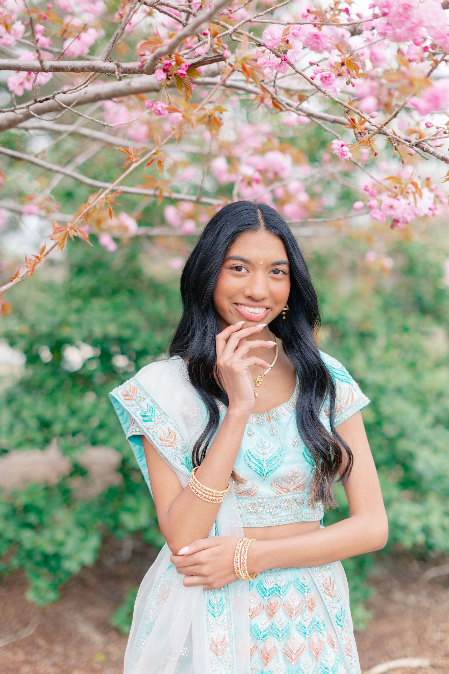 Young woman in mint embroidered lehenga smiling under pink cherry blossoms, wearing gold jewelry outdoors.