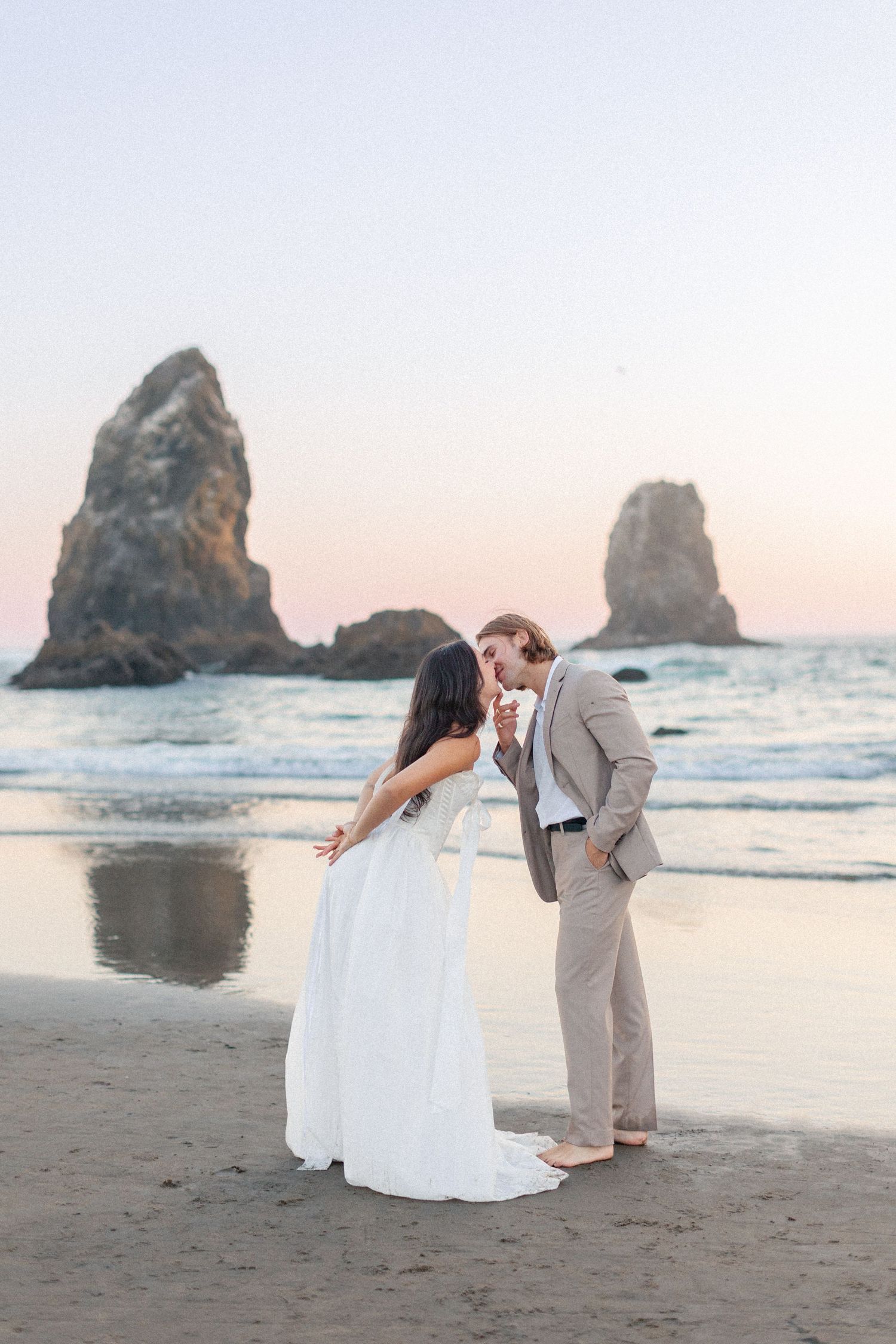 Bride and groom share a romantic moment on a sandy beach at sunset with dramatic sea stacks in the background.