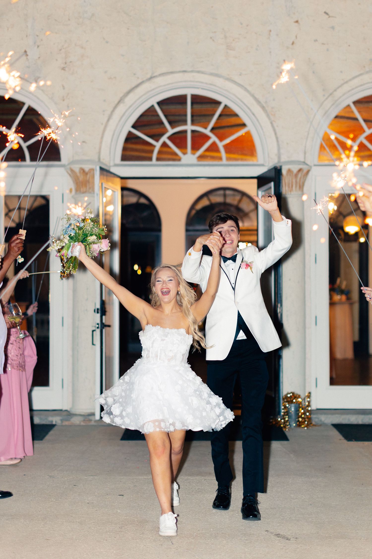 Joyful newlyweds exit wedding venue with sparklers, bride in short white dress holding bouquet, groom in white tuxedo jacket, Bella Cosa