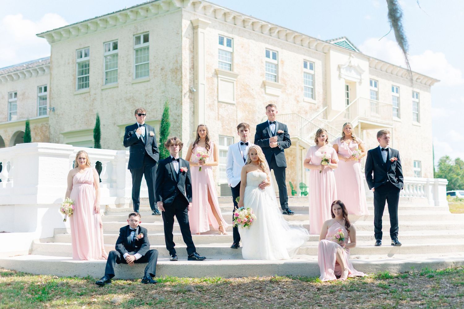 Wedding party poses on steps of elegant venue, bridesmaids in pink gowns and groomsmen in black tuxedos with bride and groom, Bella Cosa