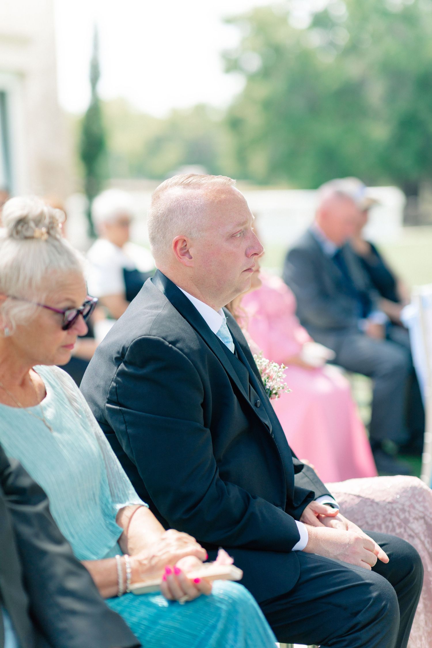 Man in black suit with boutonniere seated at outdoor wedding ceremony alongside other guests, wedding at Bella Cosa