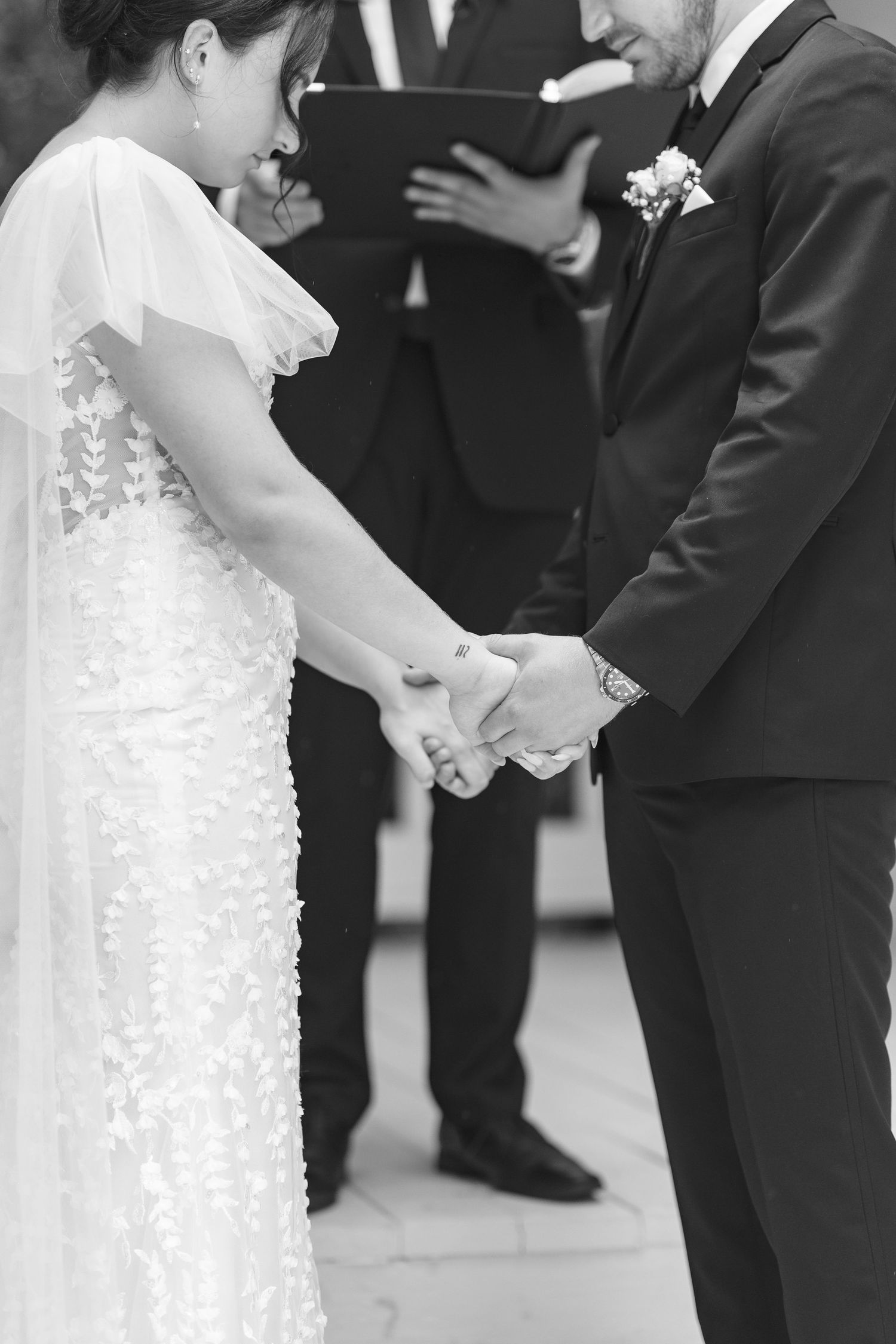 Black and white photo of bride and groom holding hands during wedding ceremony with officiant in background.