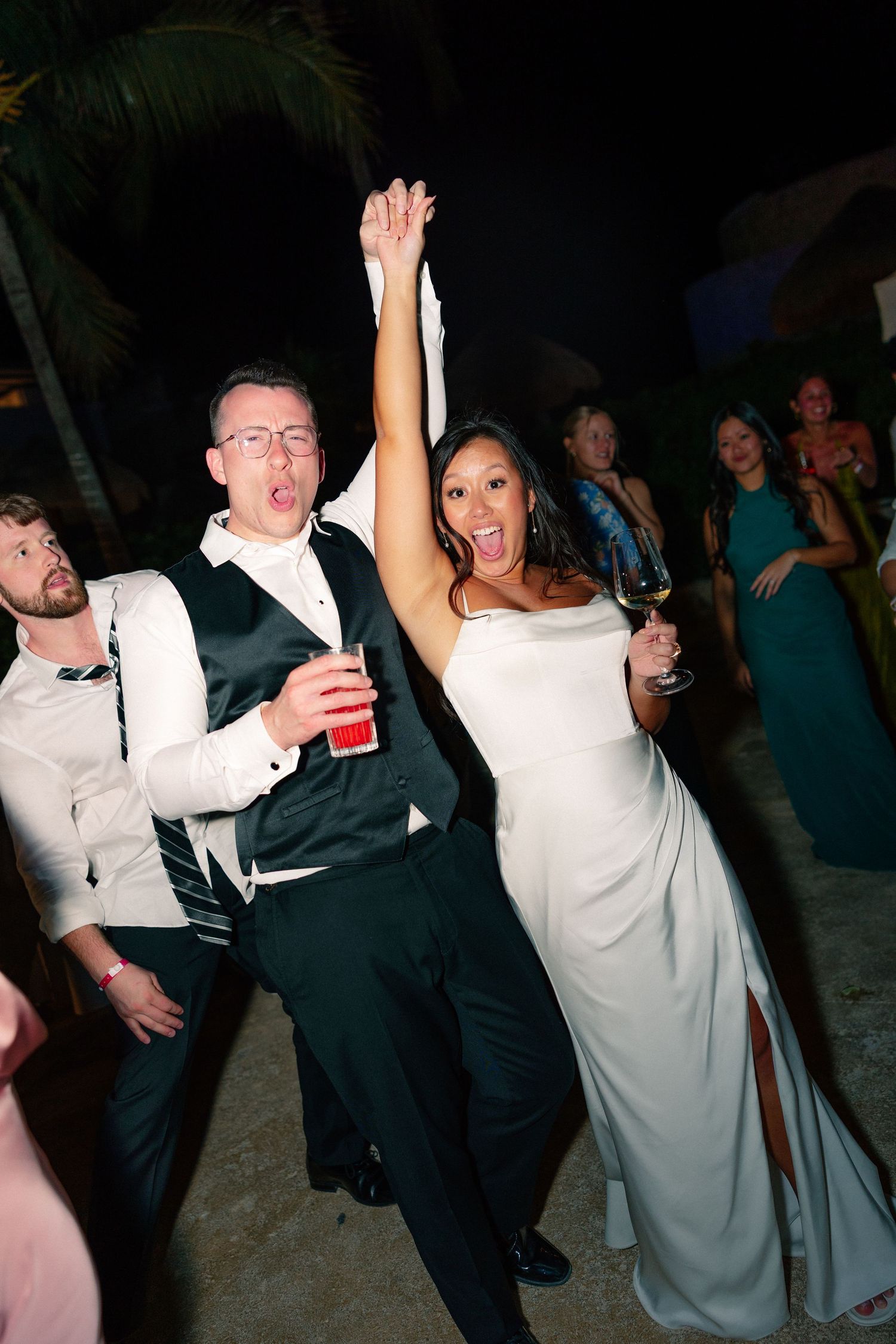 Joyful bride in white gown and groom in black suit celebrating at outdoor wedding reception, dancing with raised arms.