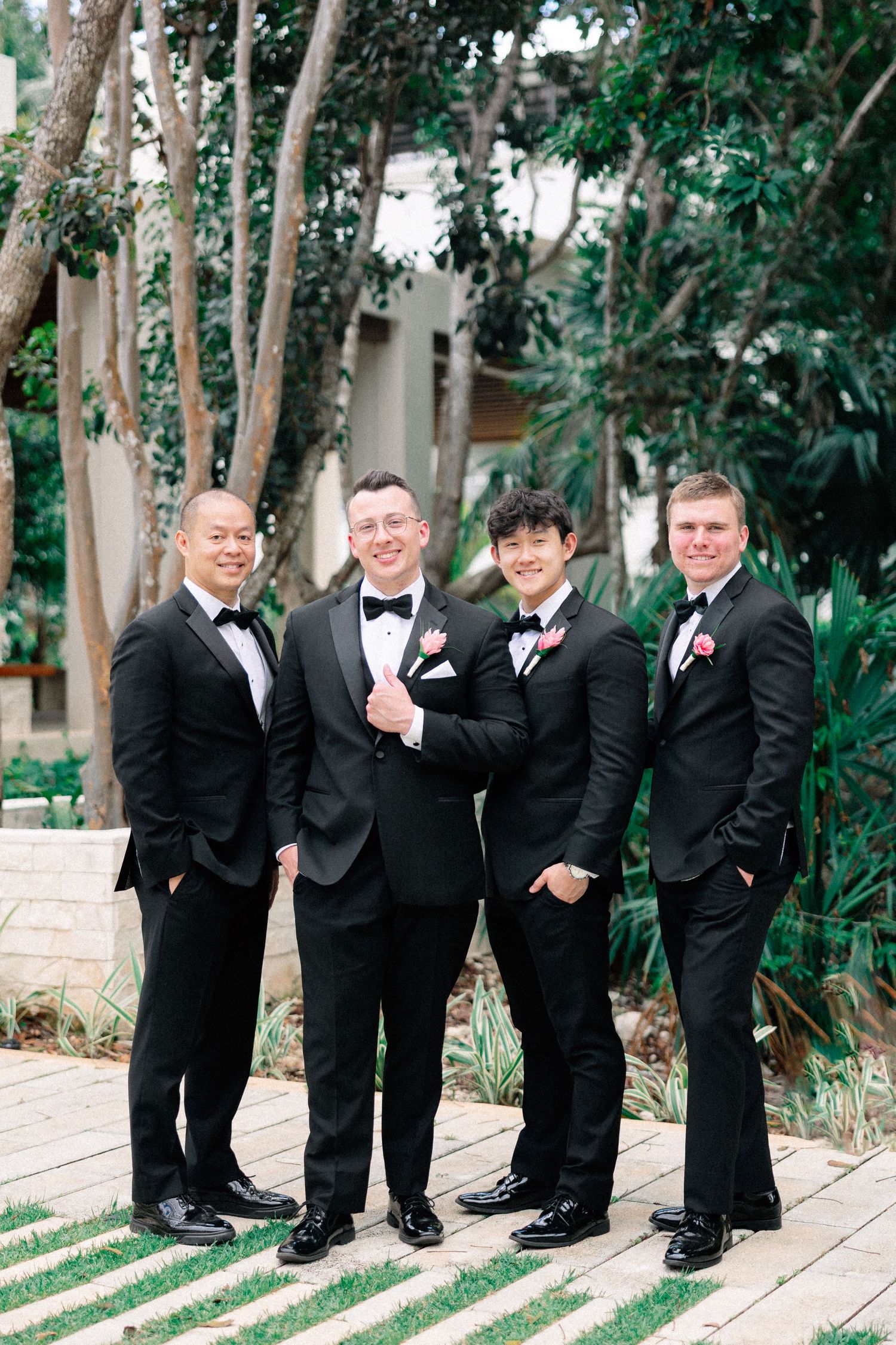 Four groomsmen in black tuxedos with bow ties and pink boutonnieres posing outdoors at a wedding venue.