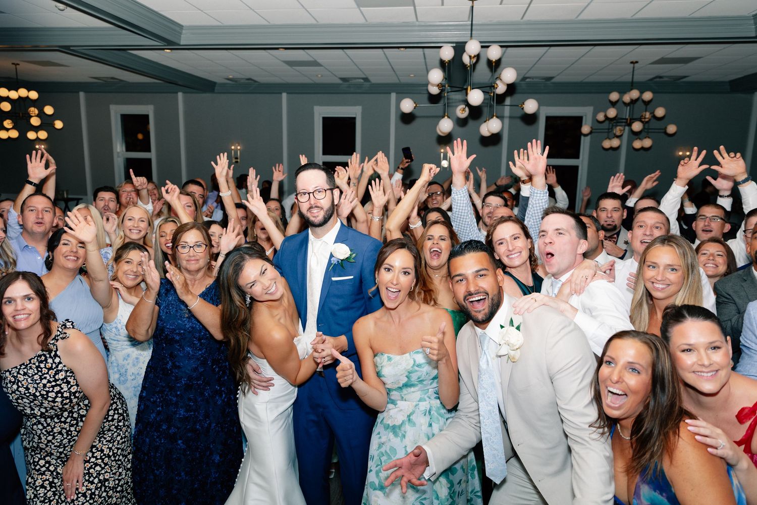 Joyful wedding reception group photo with bride and groom surrounded by cheering guests in elegant venue.