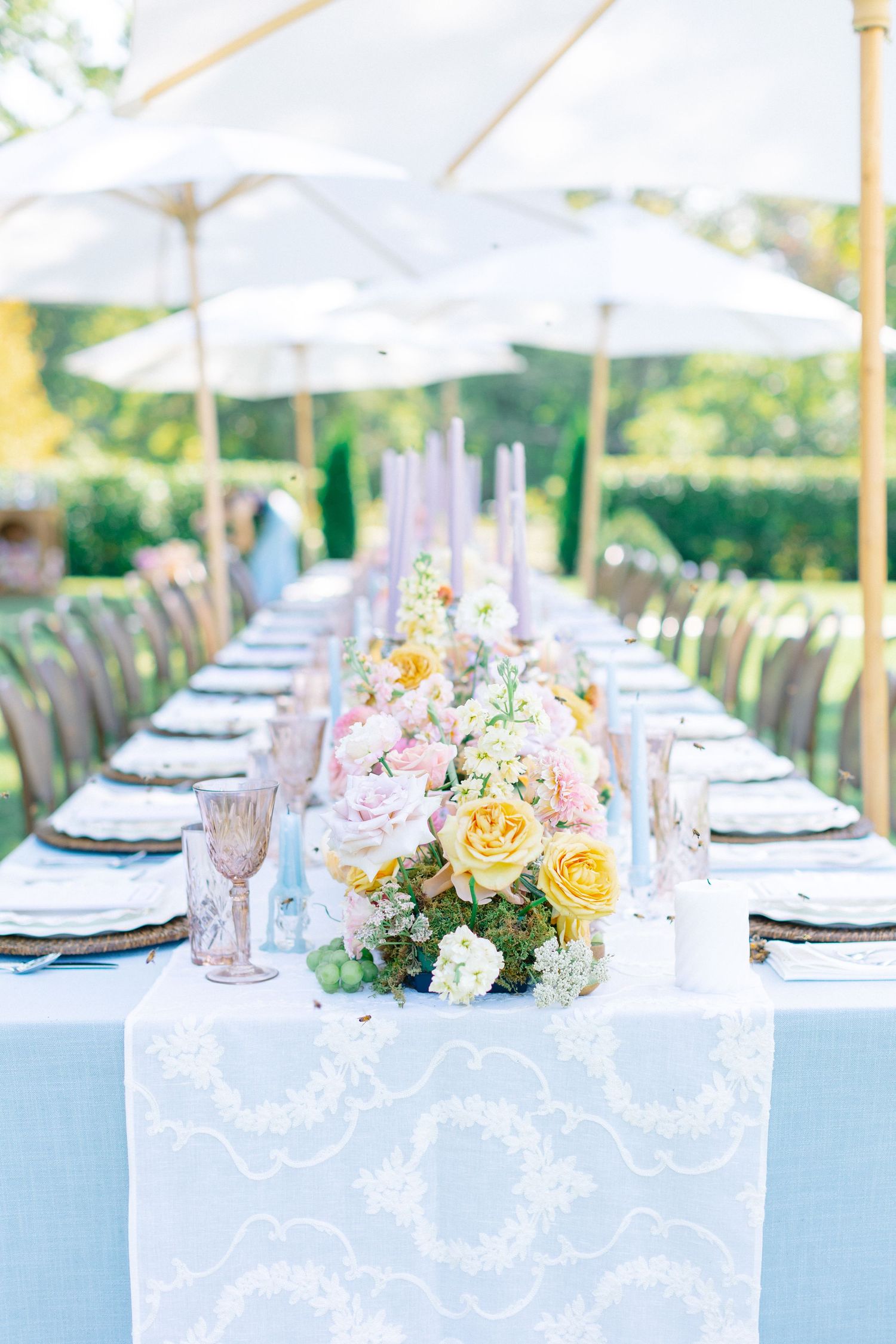 Elegant outdoor wedding reception with long table, blue runner, yellow and pink floral centerpieces, and white market umbrellas.