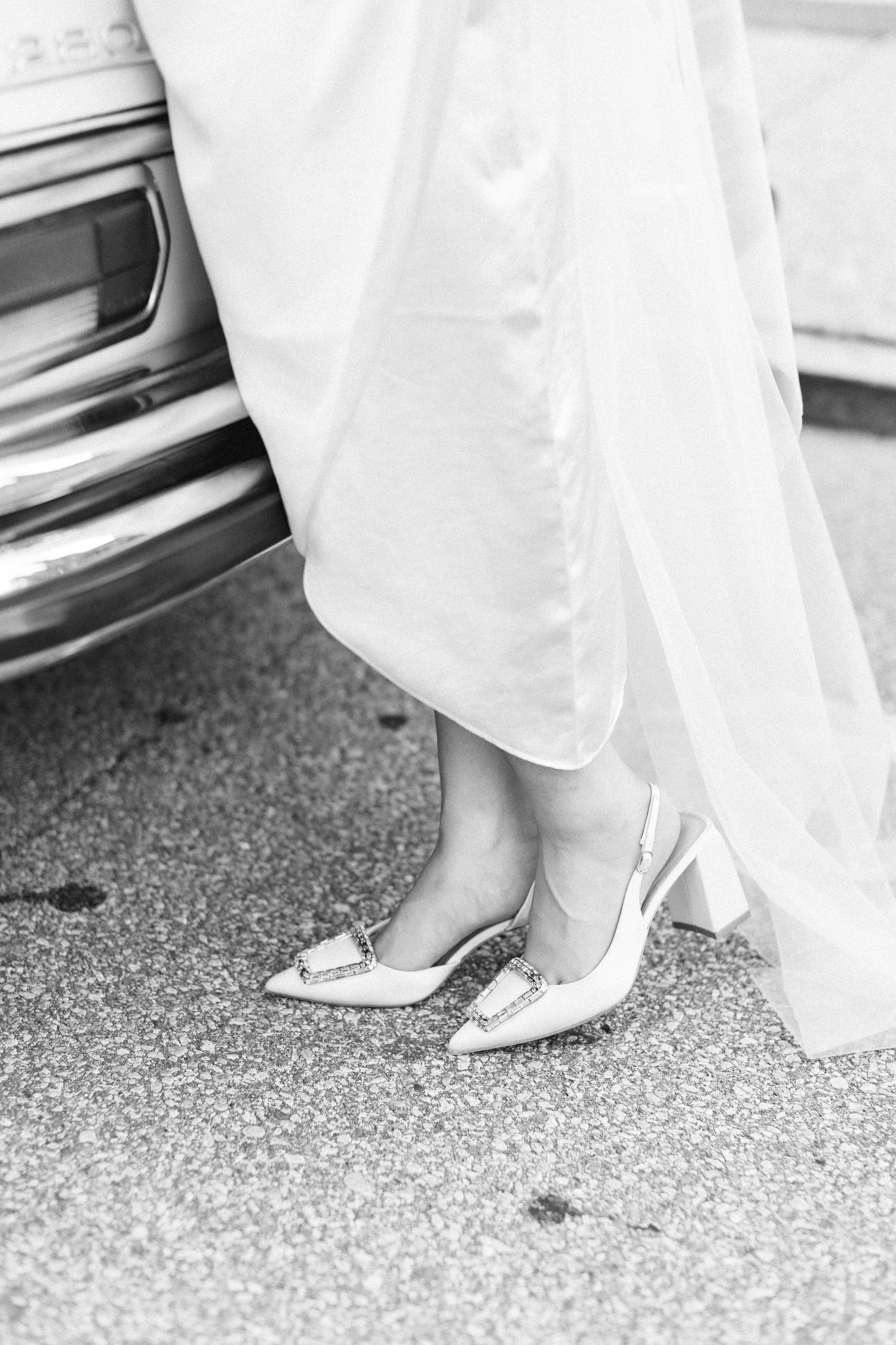 Black and white close-up of a bride wearing elegant pointed-toe slingback heels beside a vintage car.