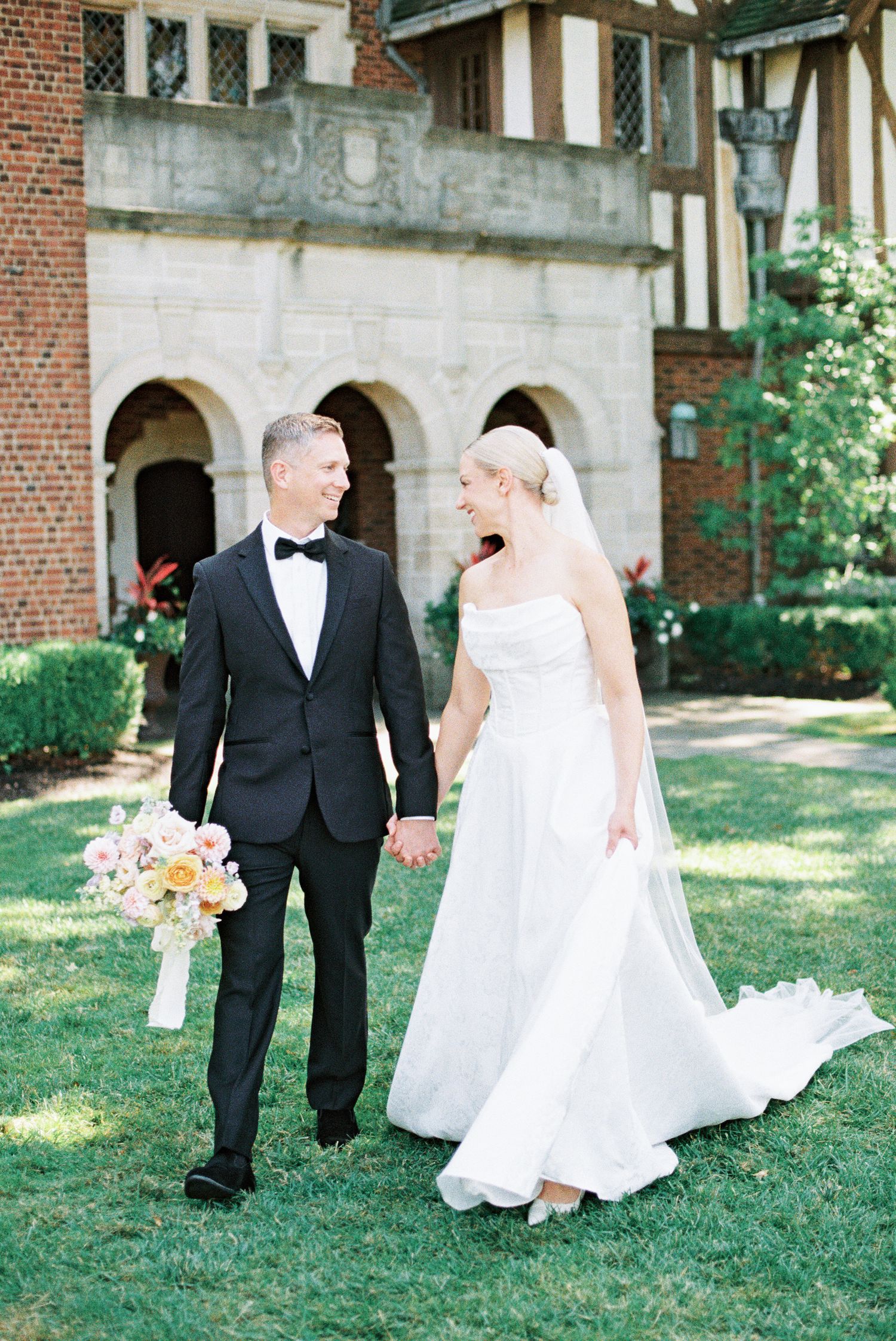 Bride and groom walk hand-in-hand laughing outside a Tudor-style brick venue, she holds a pastel bouquet.