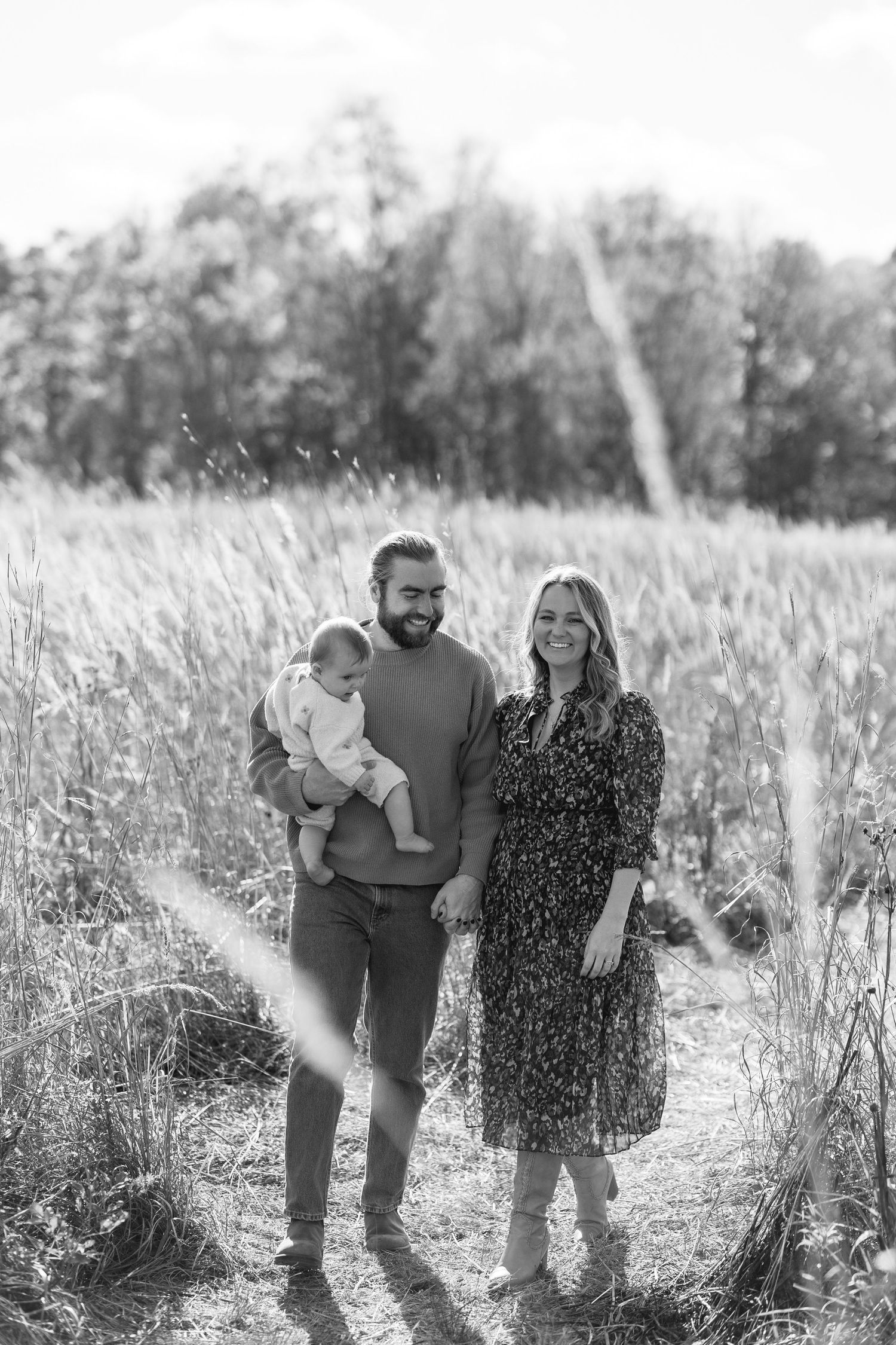A black and white portrait of a couple smiling in a golden field, the man holding their baby at French Park