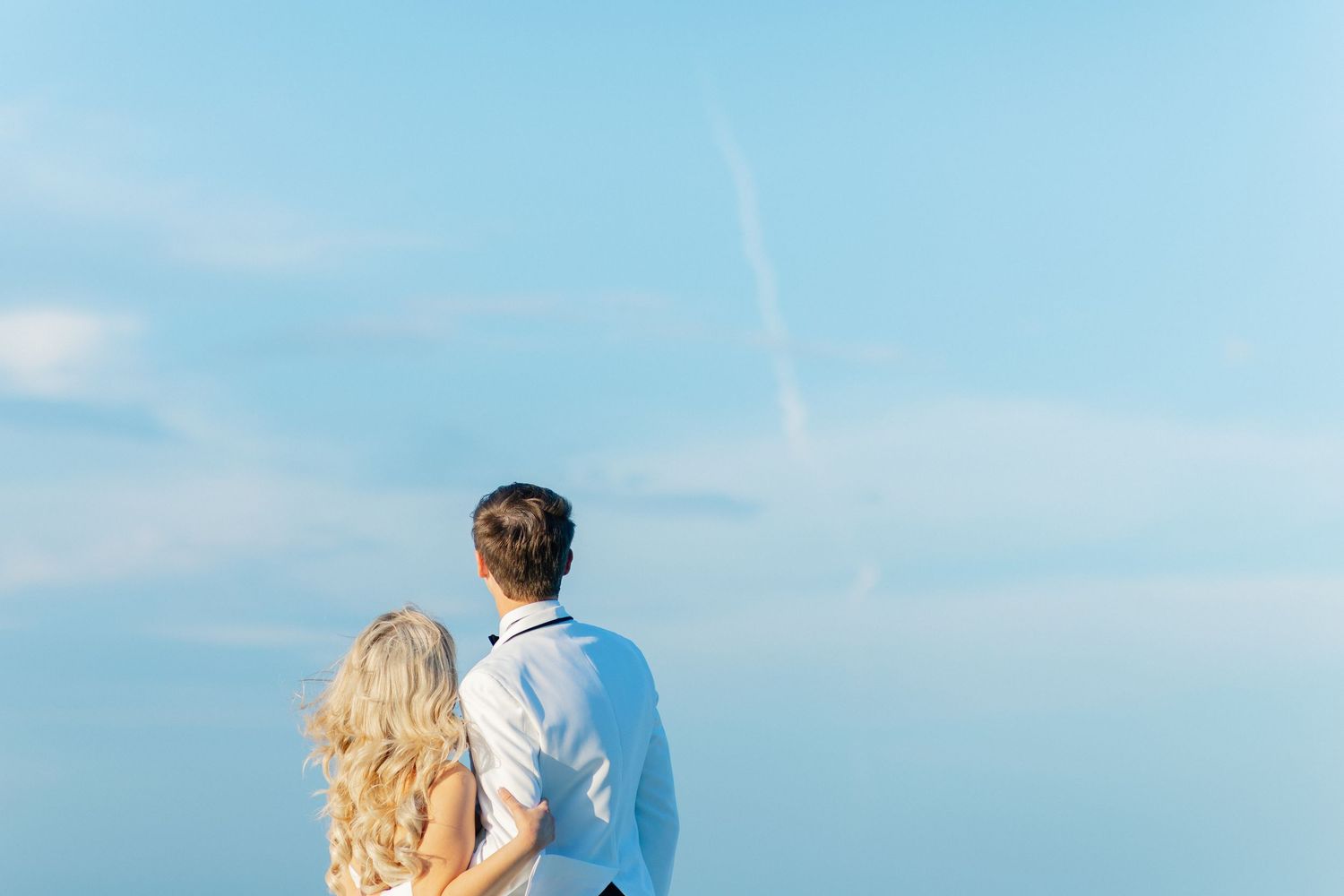 Couple embracing from behind, gazing at a clear blue sky with a faint contrail, romantic outdoor scene, wedding at Bella Cosa