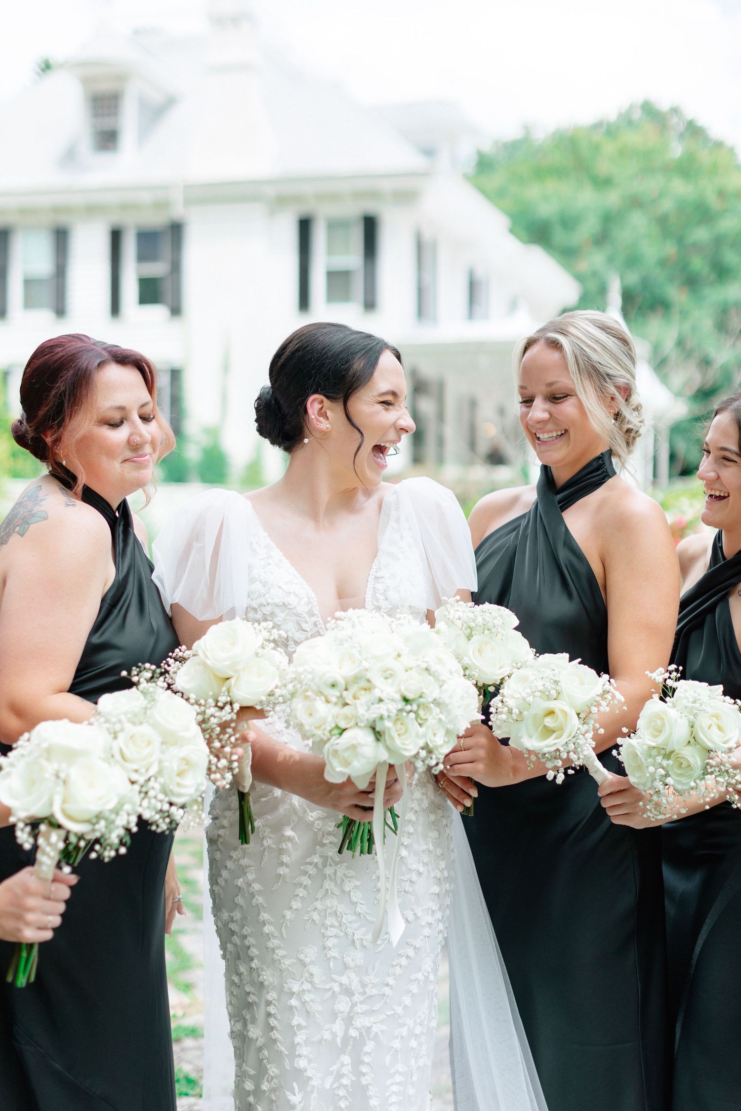Bride in white lace gown laughs with bridesmaids in black dresses holding white rose bouquets outside elegant venue.