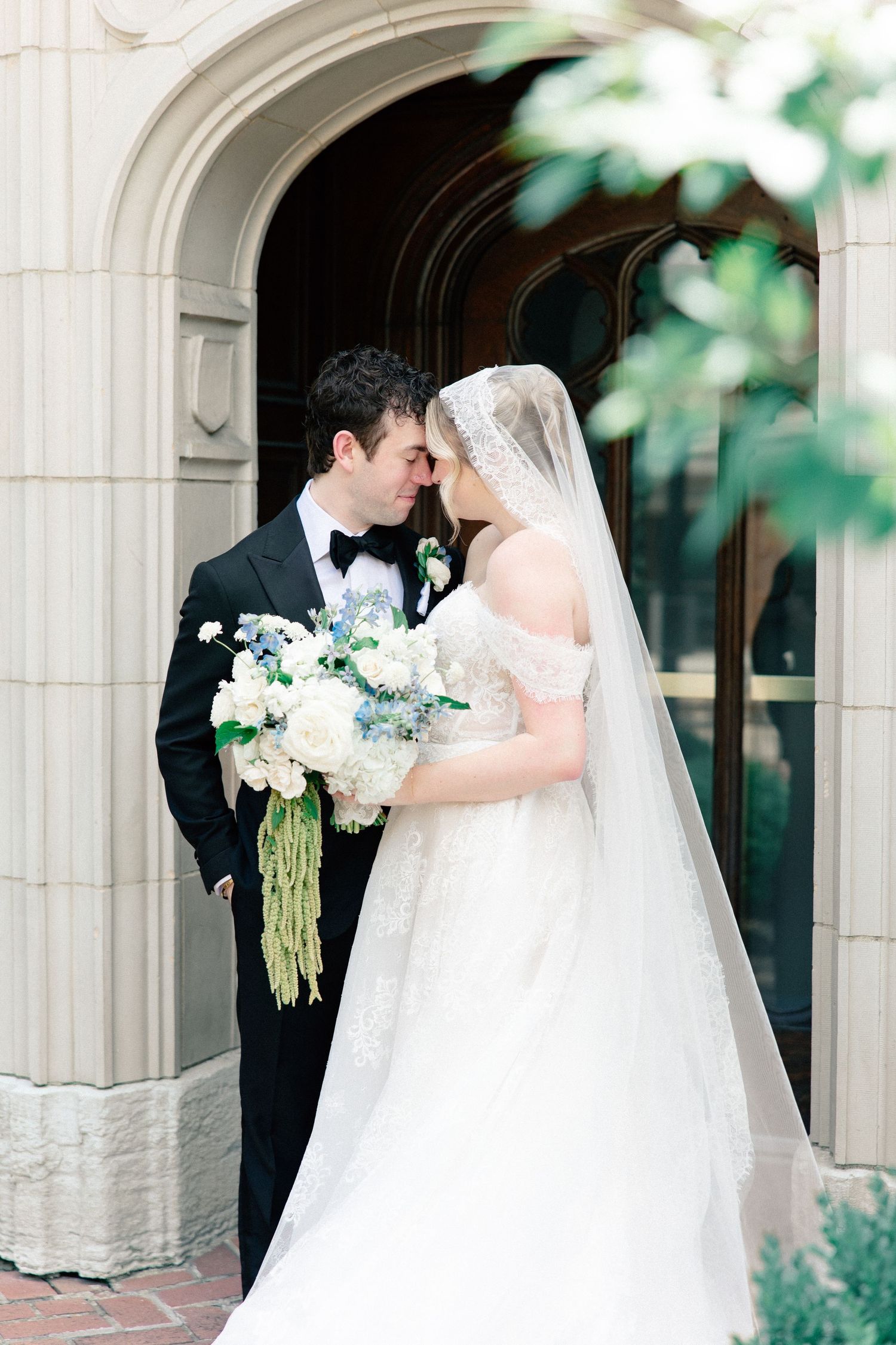 Bride and groom share a romantic moment under a stone arch, bride holding a white and blue bouquet, groom in black tuxedo.