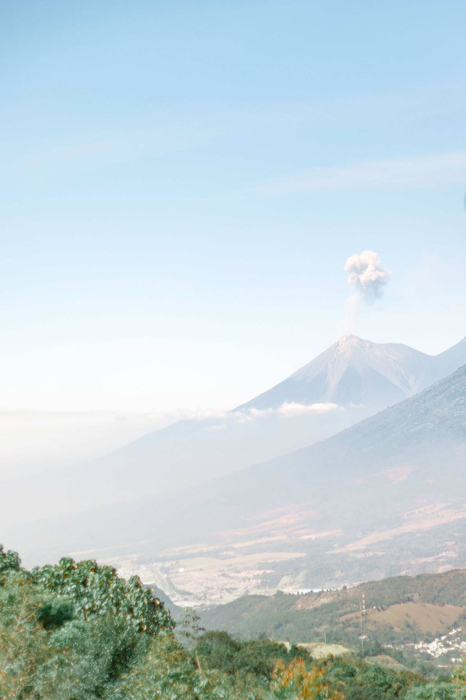 Aerial view of an active volcano emitting ash plume over a misty valley with lush green hills in Guatemala.