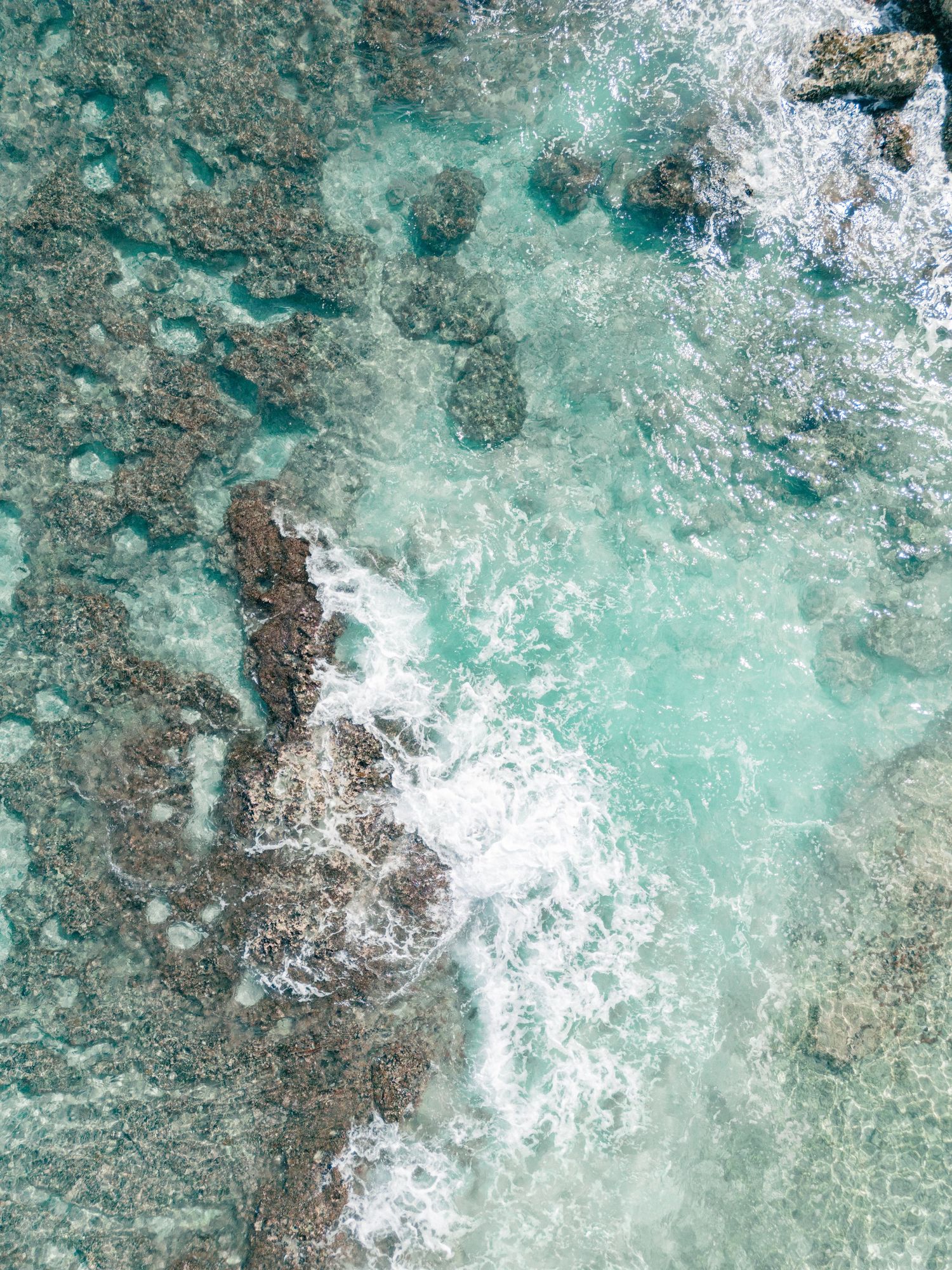 Aerial view of crystal clear turquoise ocean water washing over rocky coral reef formations near a tropical coastline.