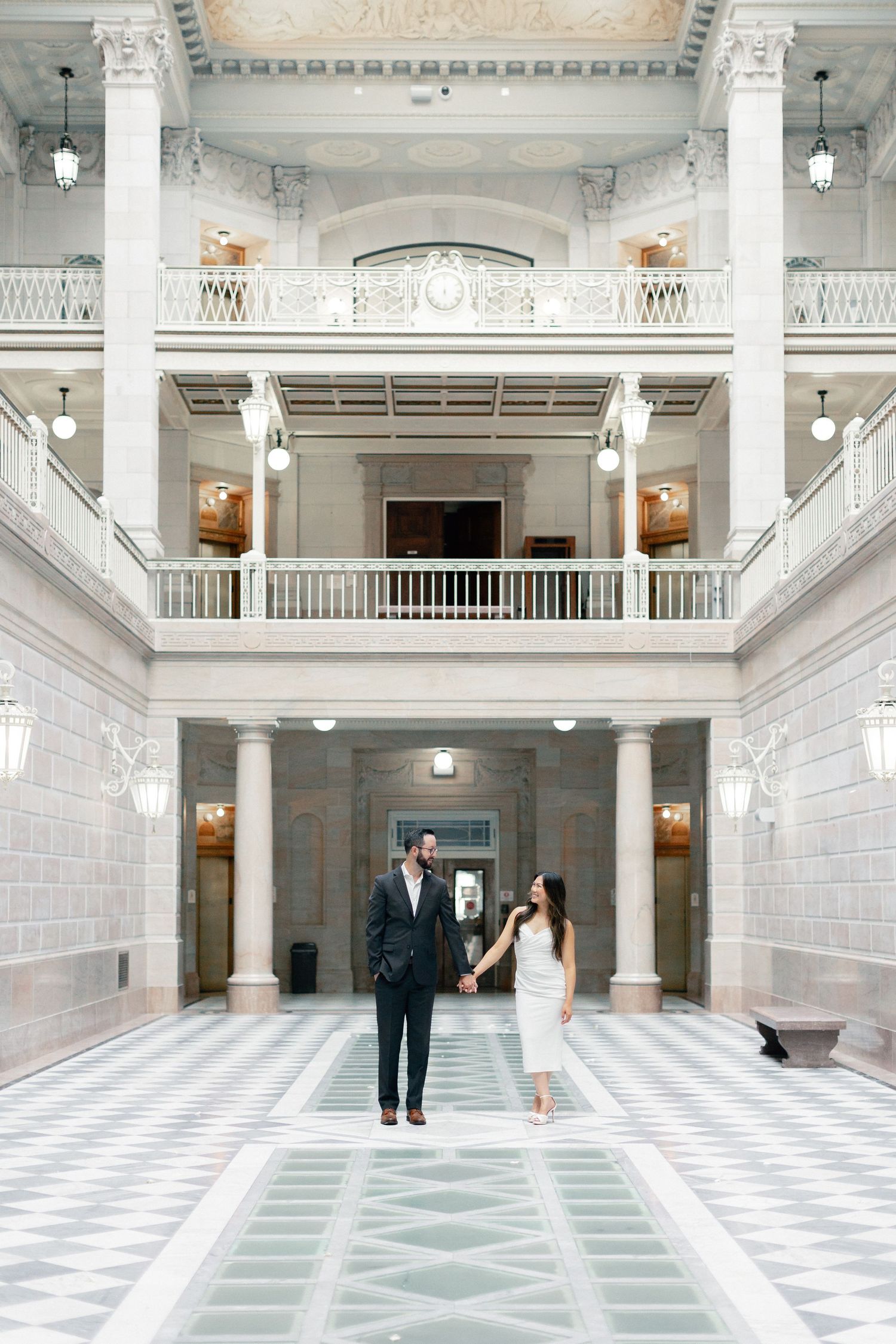 Couple holding hands in elegant marble building with grand columns and balconies.