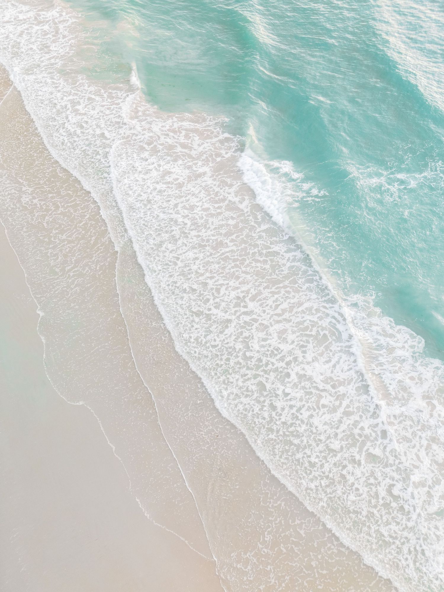 Aerial view of turquoise ocean waves gently washing onto a pristine white sandy beach.