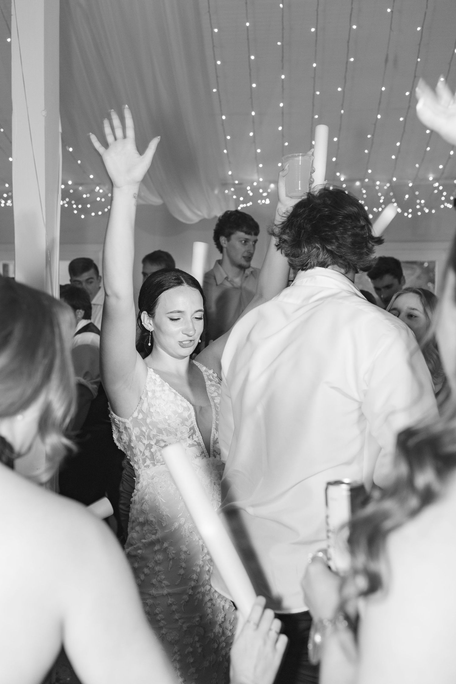 Black and white photo of a joyful woman dancing with hand raised at an elegant wedding reception under twinkling lights.