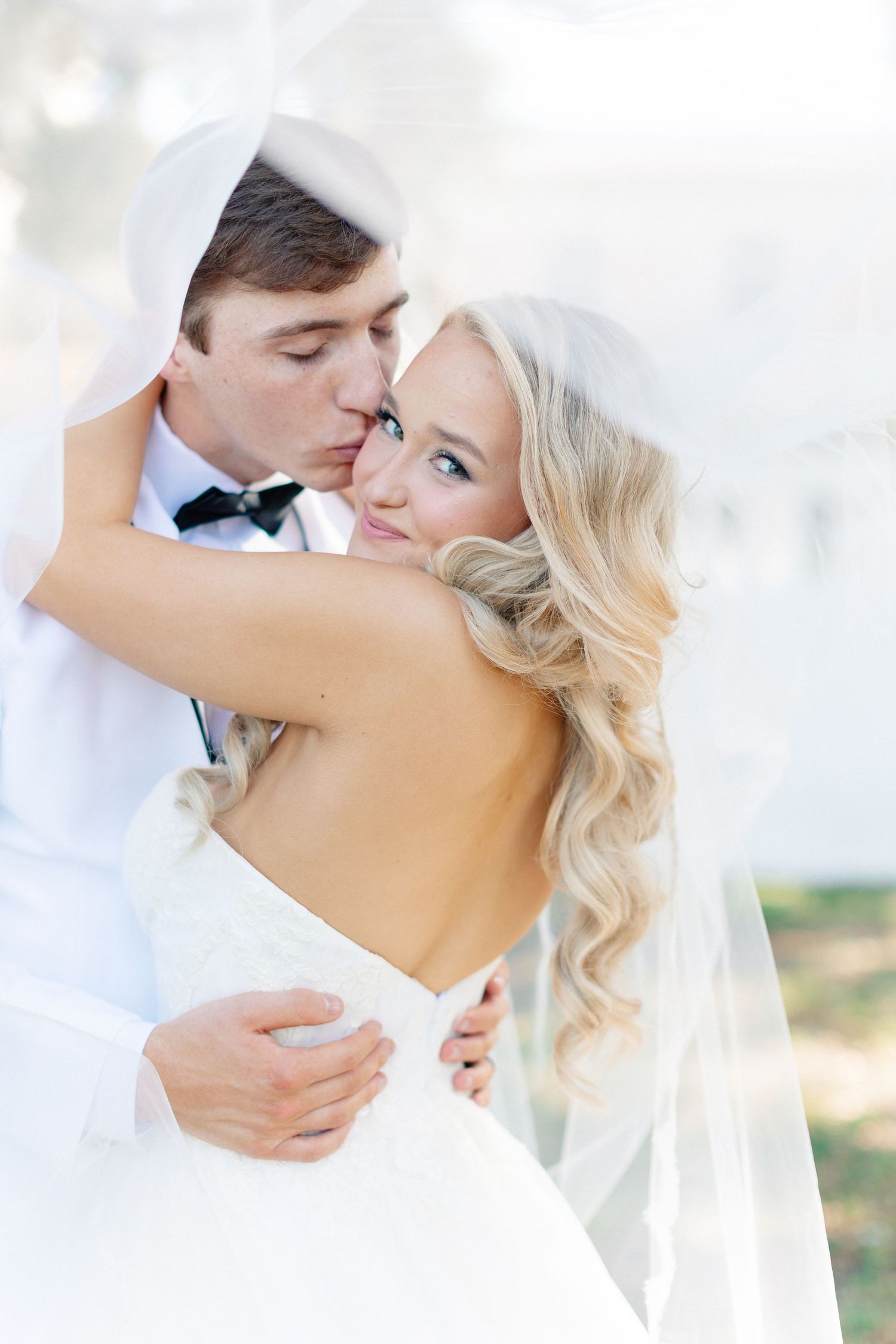 Groom kissing bride on cheek while embracing her from behind, bride in backless white gown with long veil, wedding at Bella Cosa