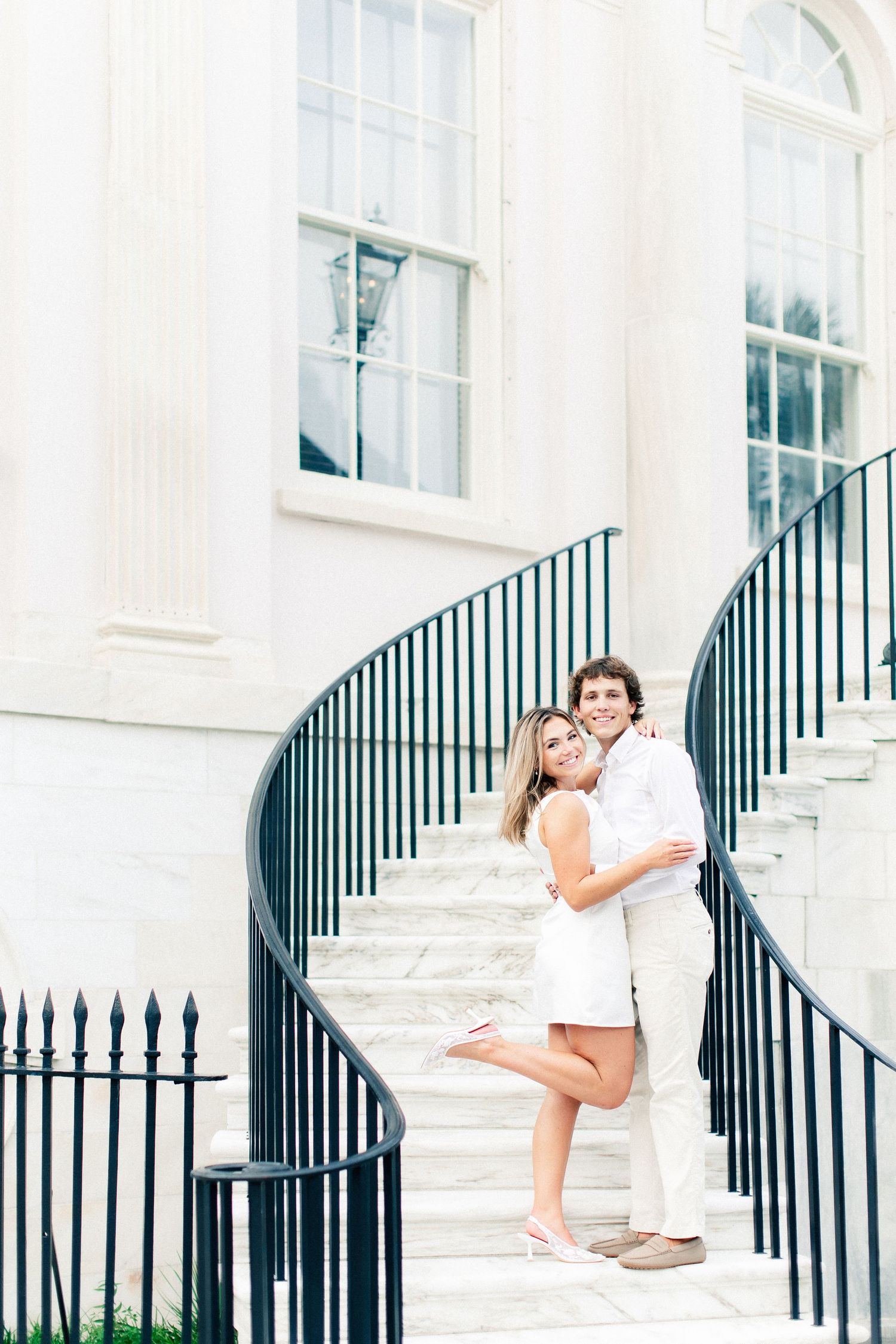 Happy couple hugging on elegant spiral staircase of a white neoclassical building, woman in white dress, man in white outfit.