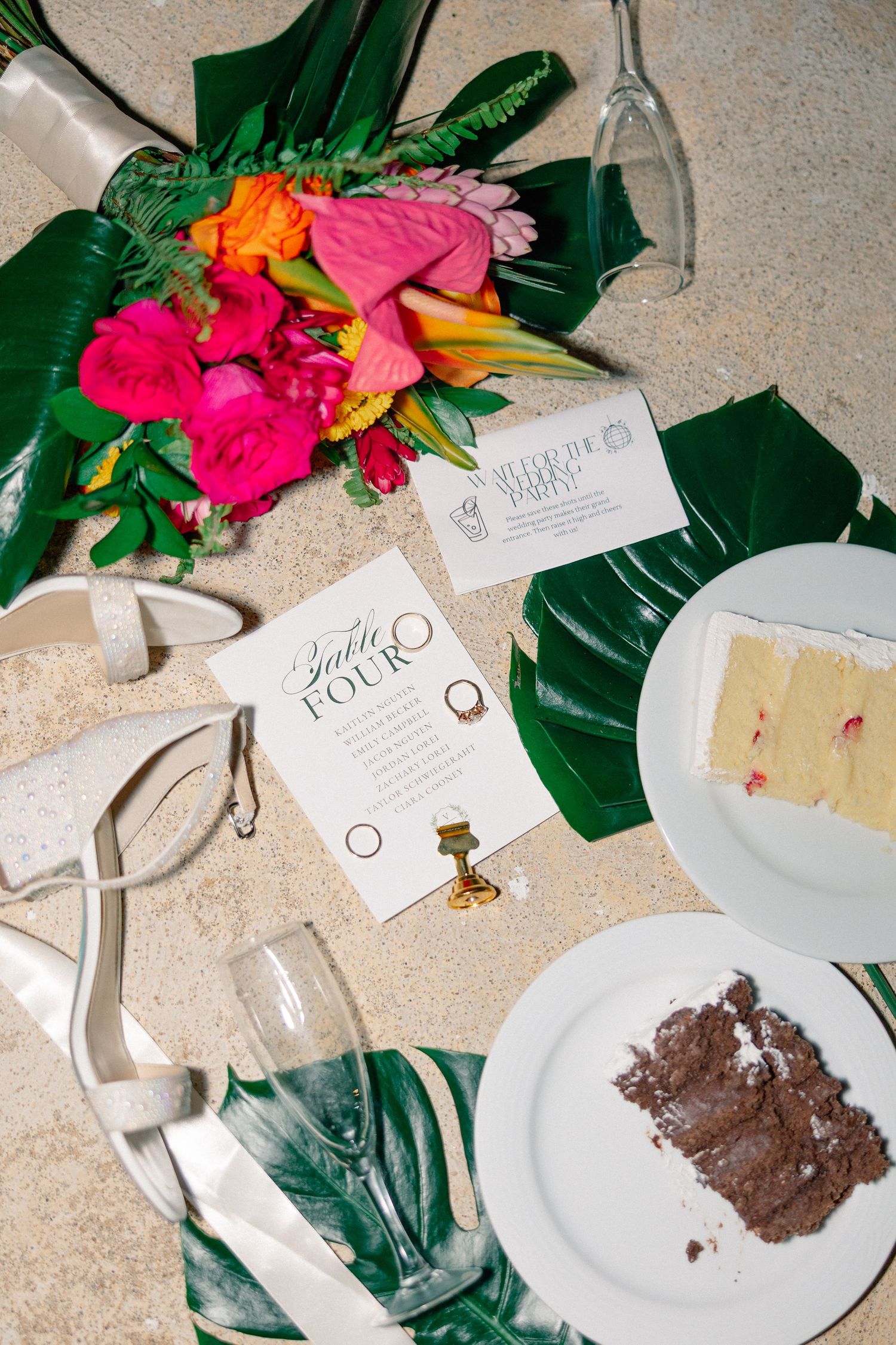 Tropical wedding flat lay with vibrant bouquet, white heels, cake slices, monstera leaves, and elegant stationery on sandy surface.