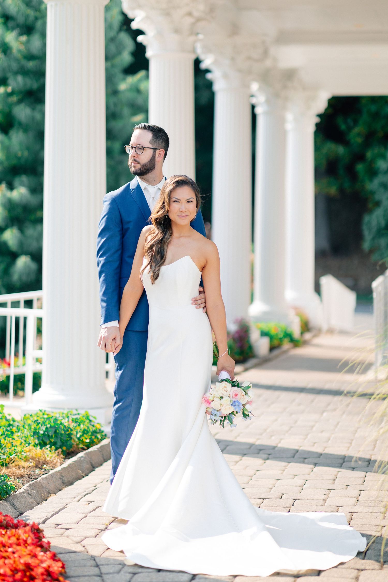 Bride in white gown holding bouquet stands with groom in blue suit among white columns at outdoor wedding venue.