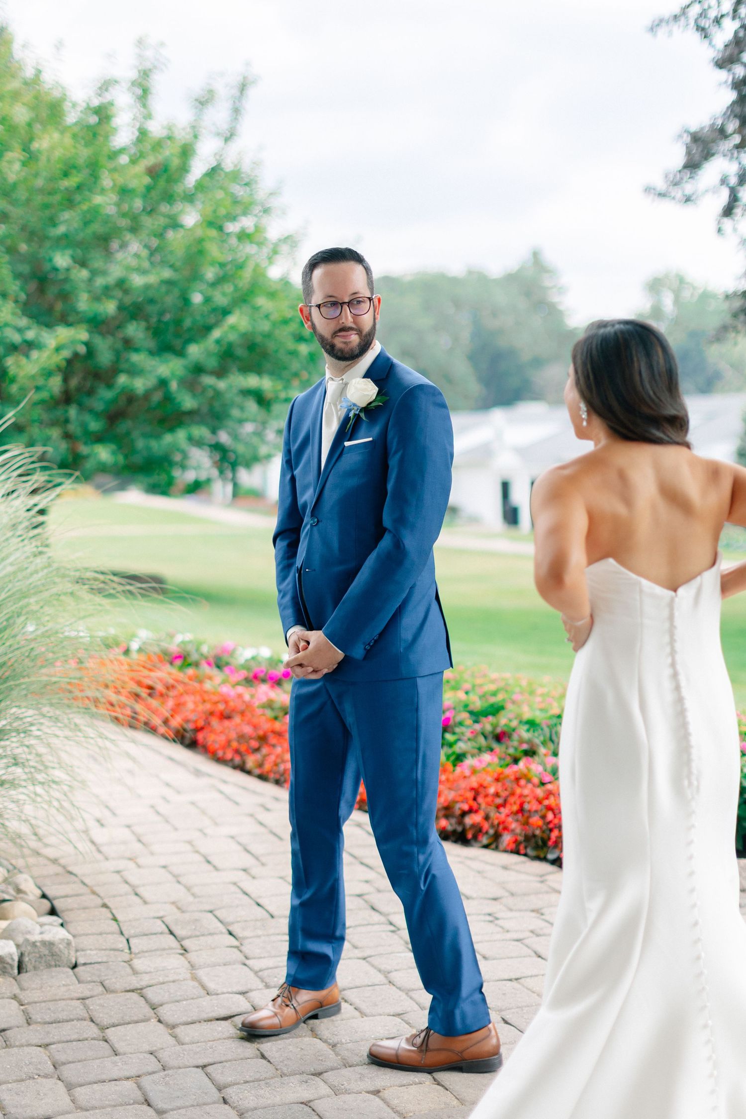 Groom in navy blue suit turns to see bride in white strapless gown during first look at outdoor wedding venue.