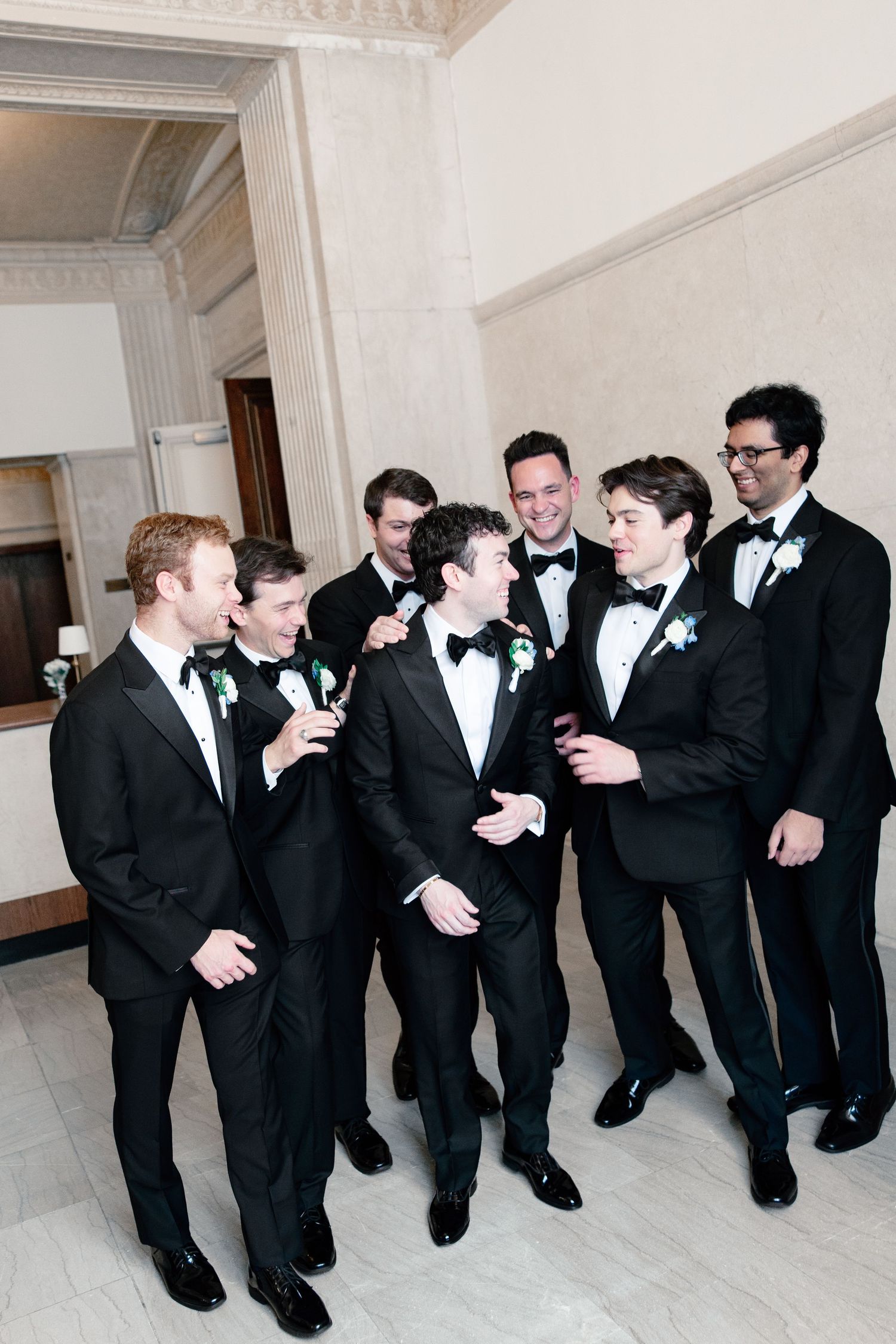 Seven groomsmen in black tuxedos with boutonnieres laughing together in an elegant marble venue.