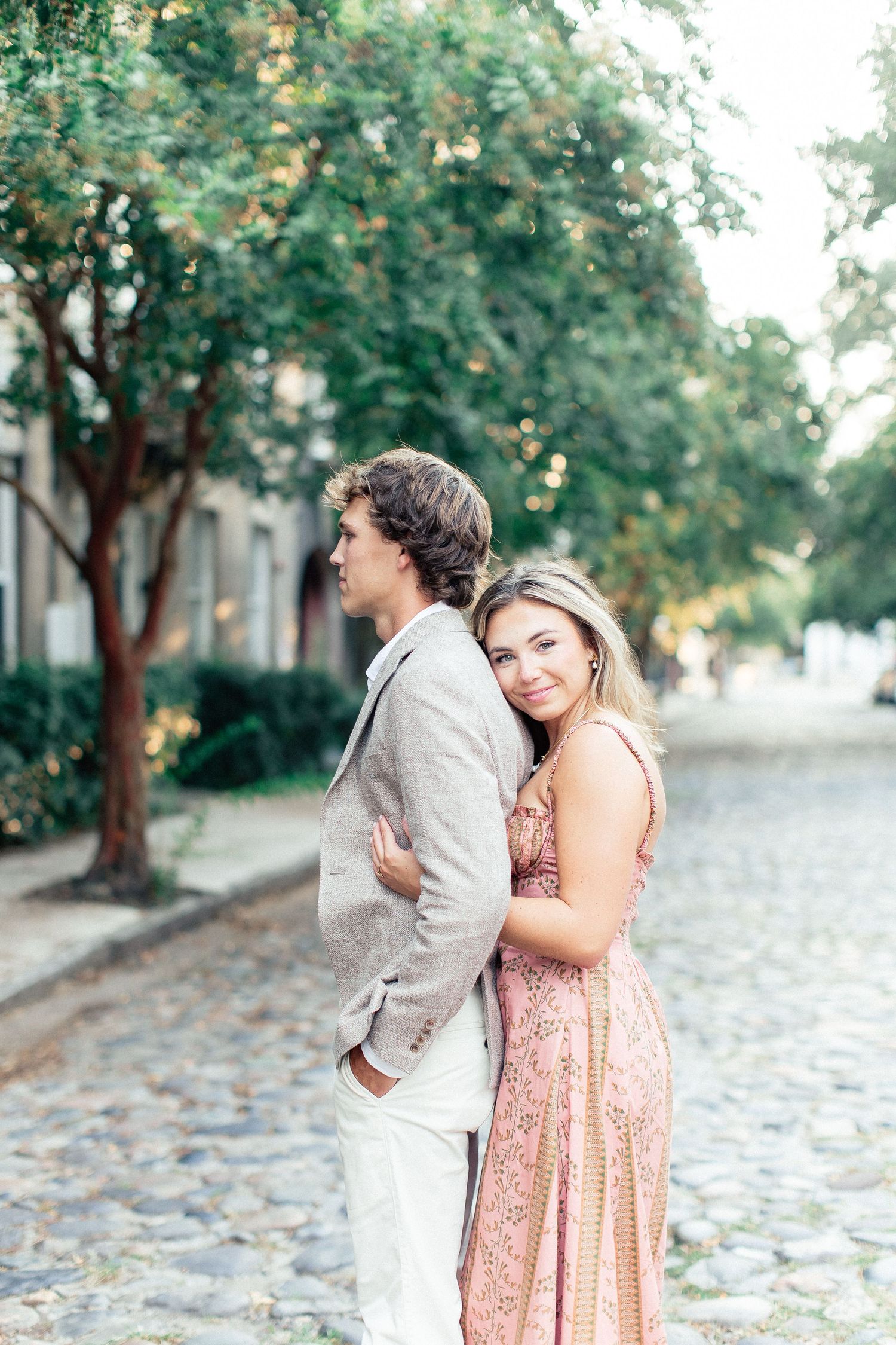 Couple embracing on cobblestone street, woman in floral dress smiling at camera, man in linen suit, surrounded by green trees.