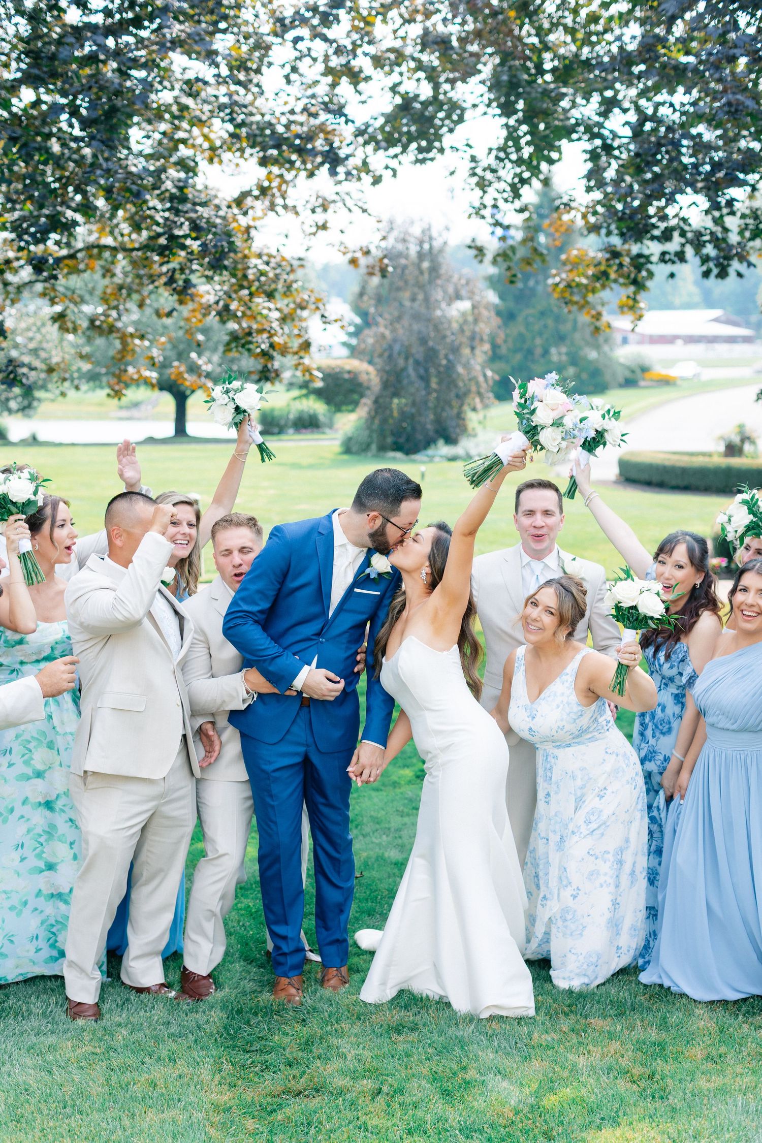 A joyful wedding party celebrates as the bride and groom share a kiss outdoors, surrounded by cheering bridesmaids and groomsmen.