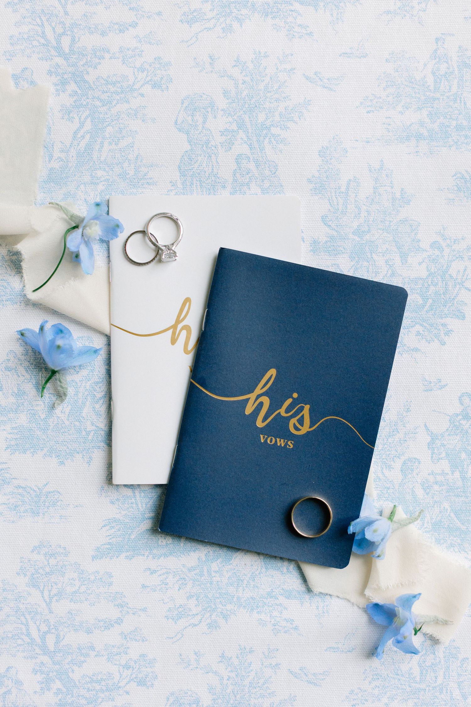 Navy and white vow books with gold script, wedding rings, and blue flowers on a blue toile background.