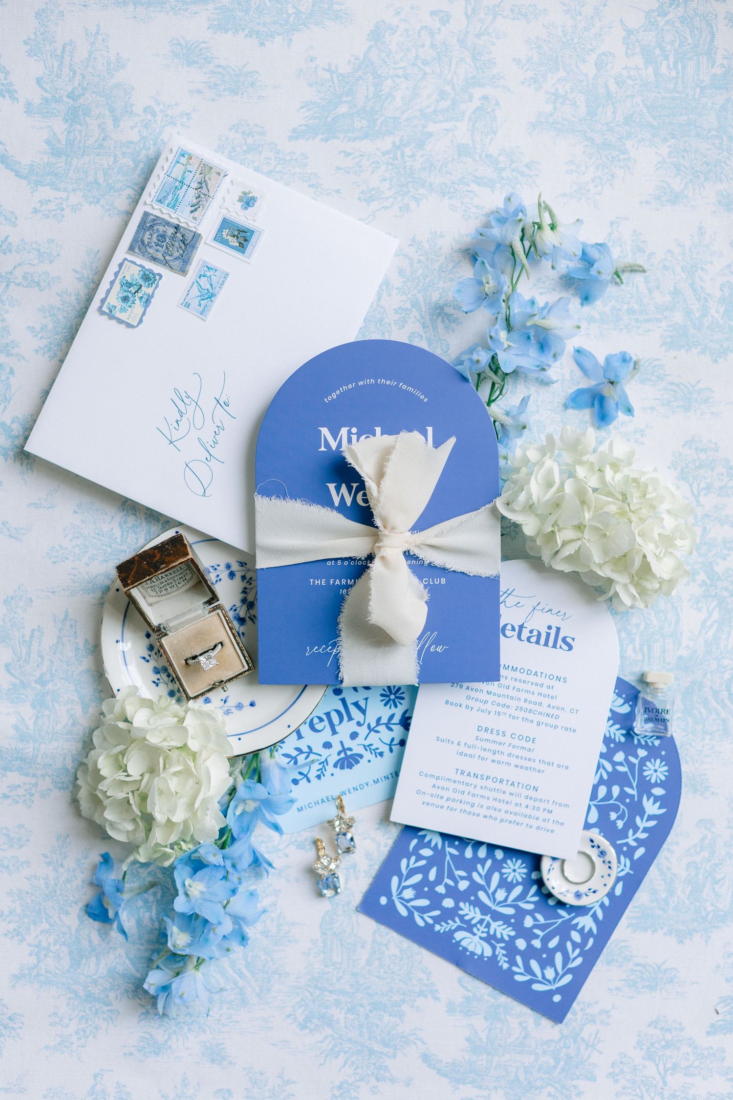 Blue and white wedding invitation suite with floral accents, ribbon-tied card, rings, and hydrangeas on toile background.