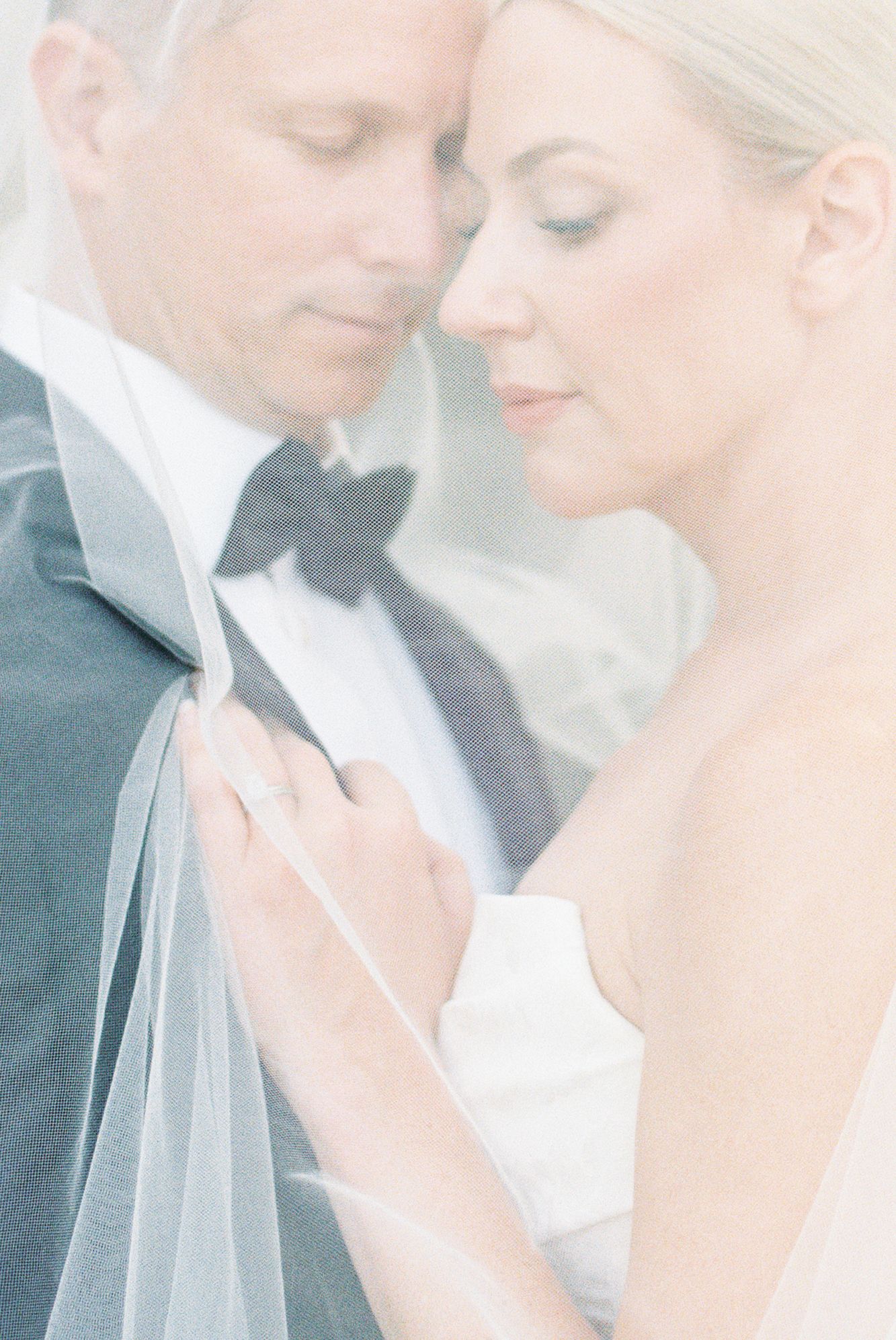 Elegant bride and groom in soft focus, bride in strapless gown holding veil, groom in black tuxedo, intimate wedding portrait.