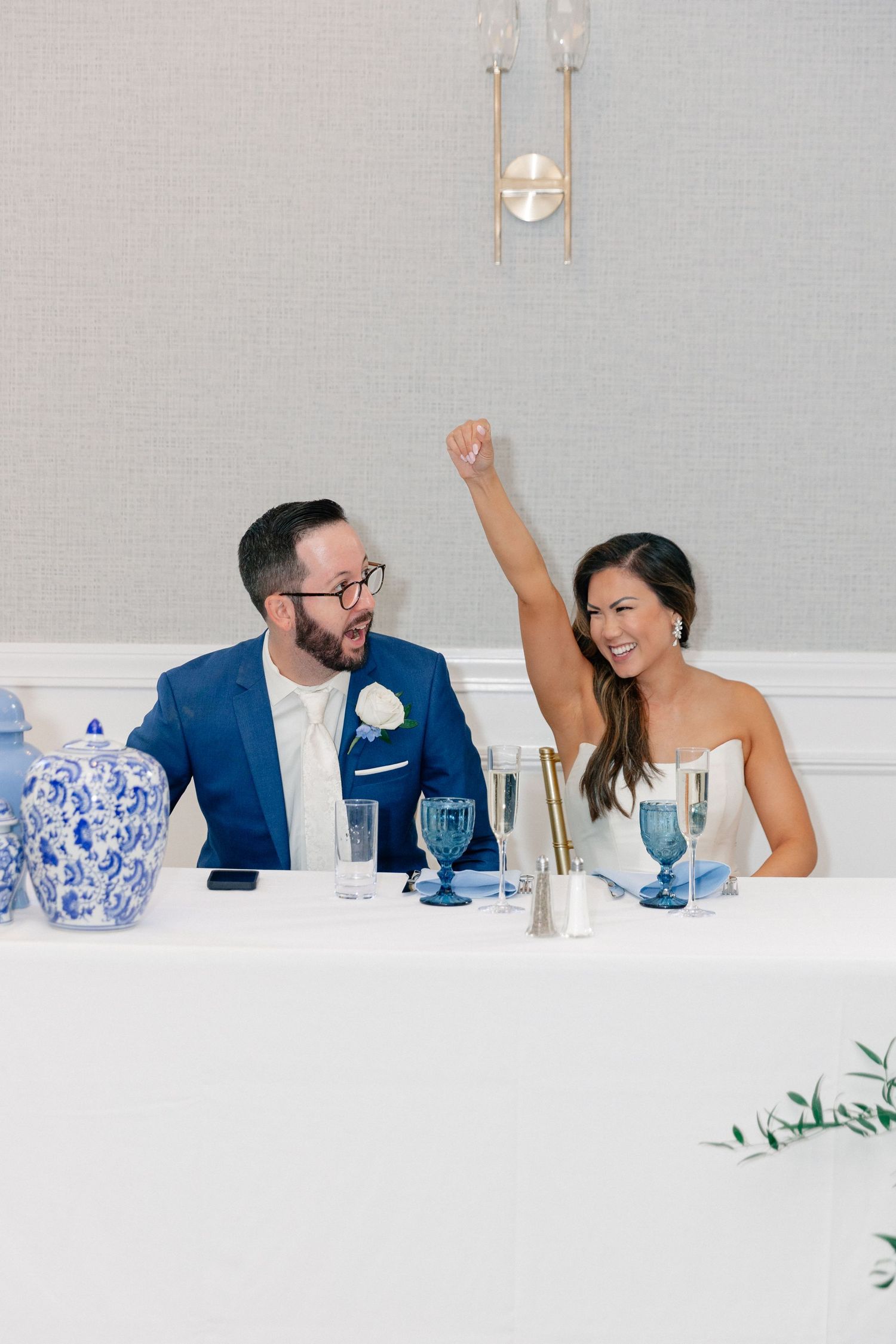 Bride raises fist in celebration beside groom in blue suit at elegant wedding reception table with blue glassware.