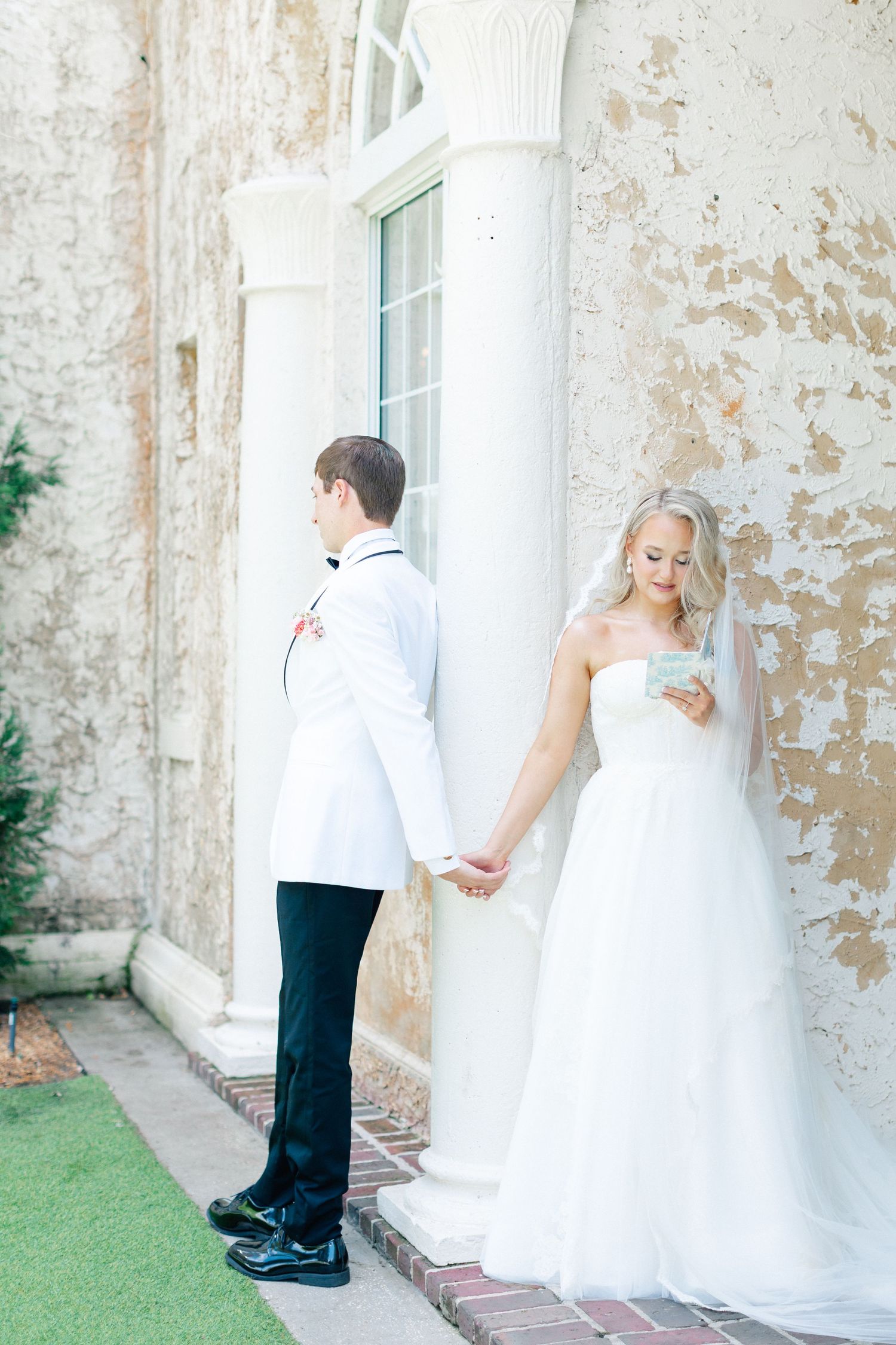 Bride and groom hold hands around a white column, she reads a note in her wedding gown and veil, wedding at Bella Cosa