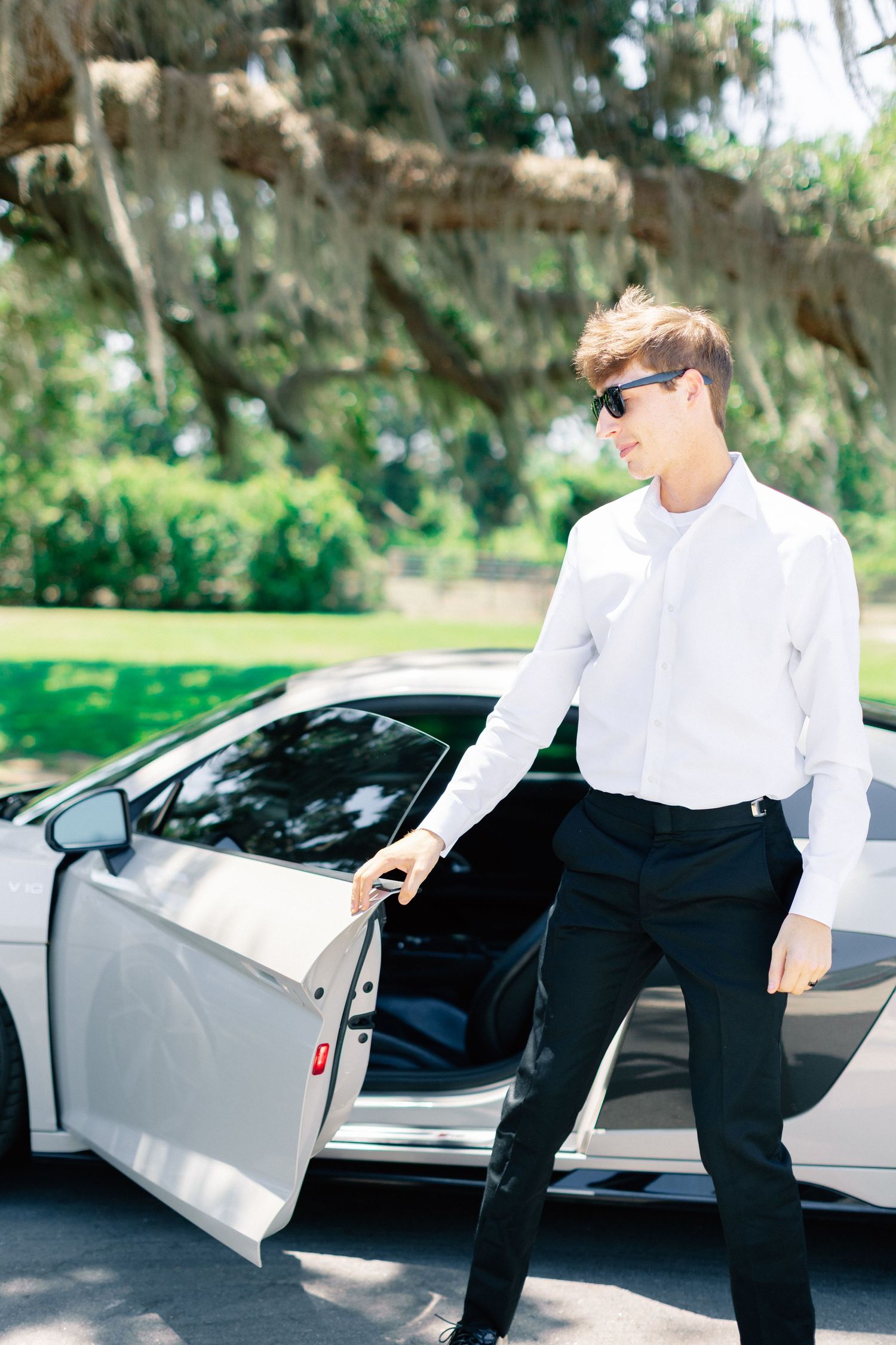 Young man in white shirt and black pants with sunglasses exiting a white luxury sports car outdoors, wedding at Bella Cosa